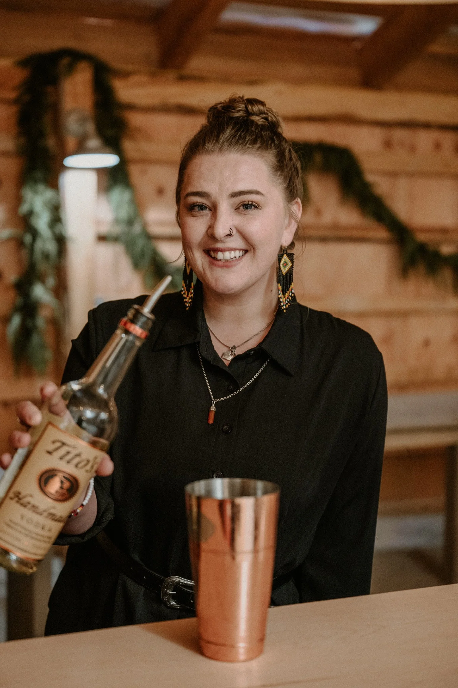 A young woman with braided hair, wearing large beaded earrings, smiling, holding a bottle of liquor behind a bar, in a wooden interior setting with hanging plant decorations.
