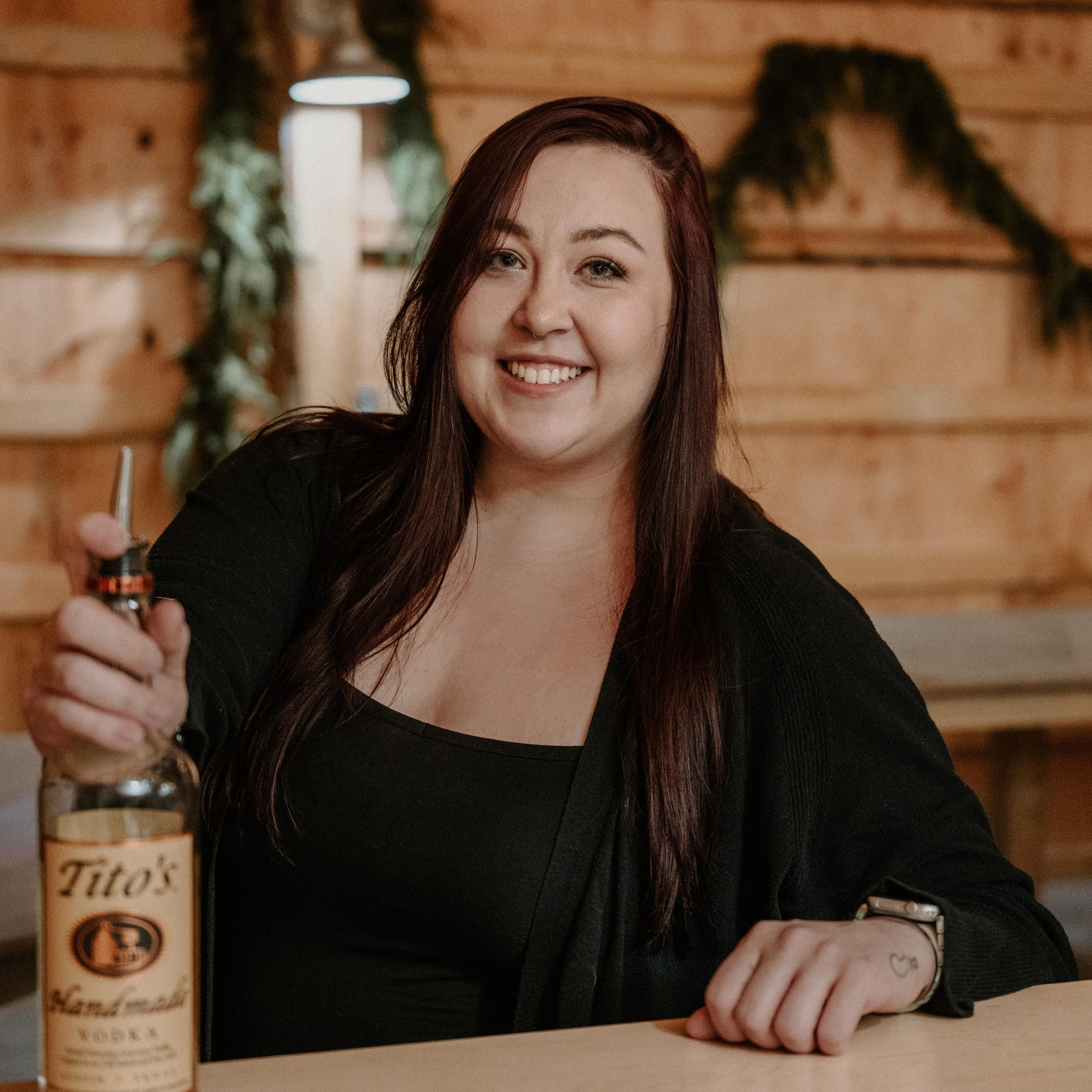 A woman with long brown hair, smiling, taking a selfie with a smartphone. She has a septum piercing and is wearing a black top. The background features a wooden wall with some greenery and she is holding a Tito's vodka liquor bottle.
