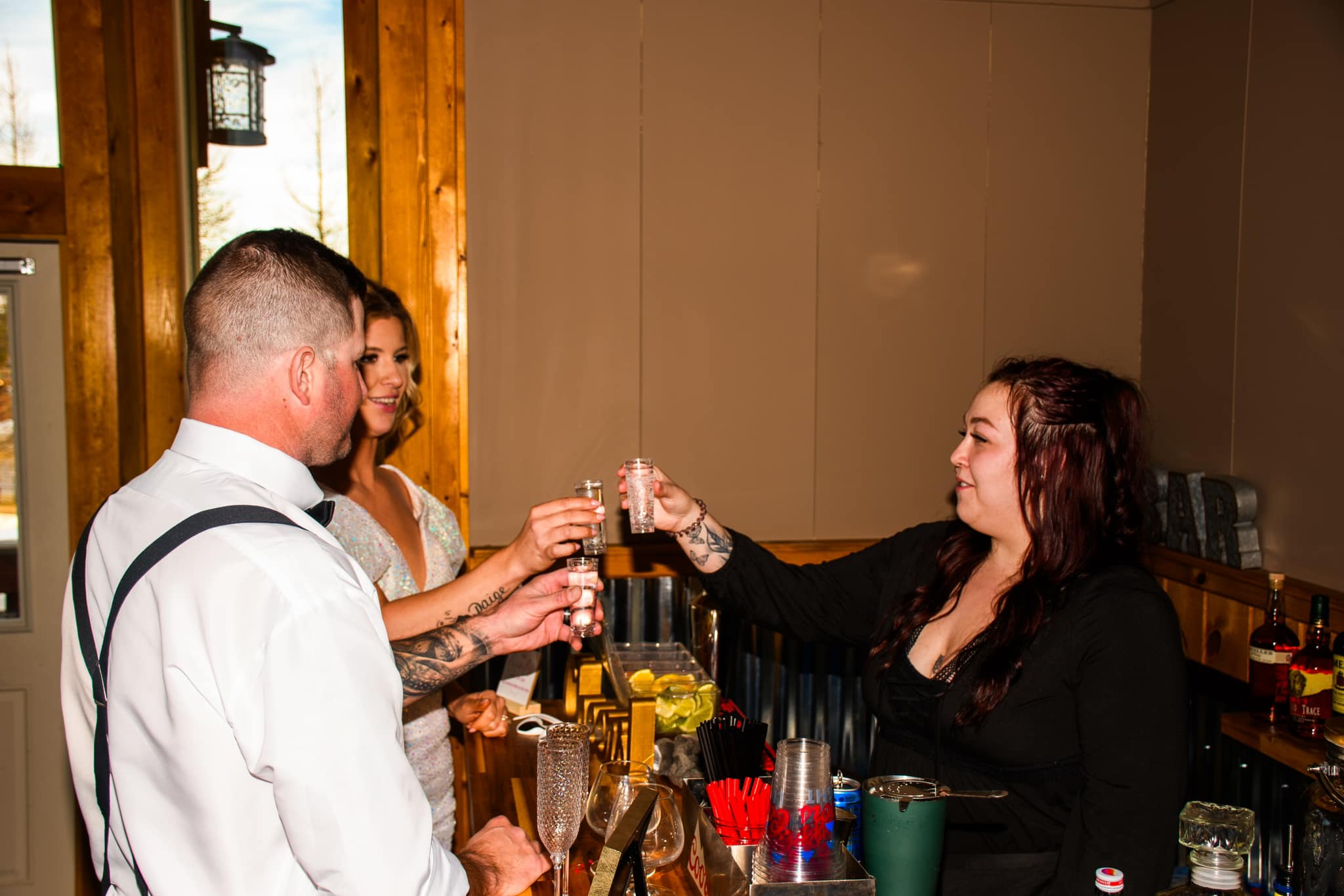 Four people raising shot glasses for a toast inside a rustic restaurant or bar with wooden walls and a window showing trees outside.