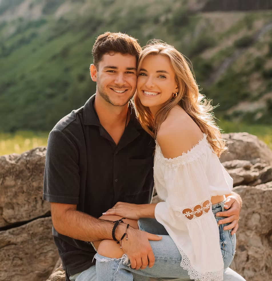 A smiling young couple sitting outdoors on rocks in a green, mountainous area.
