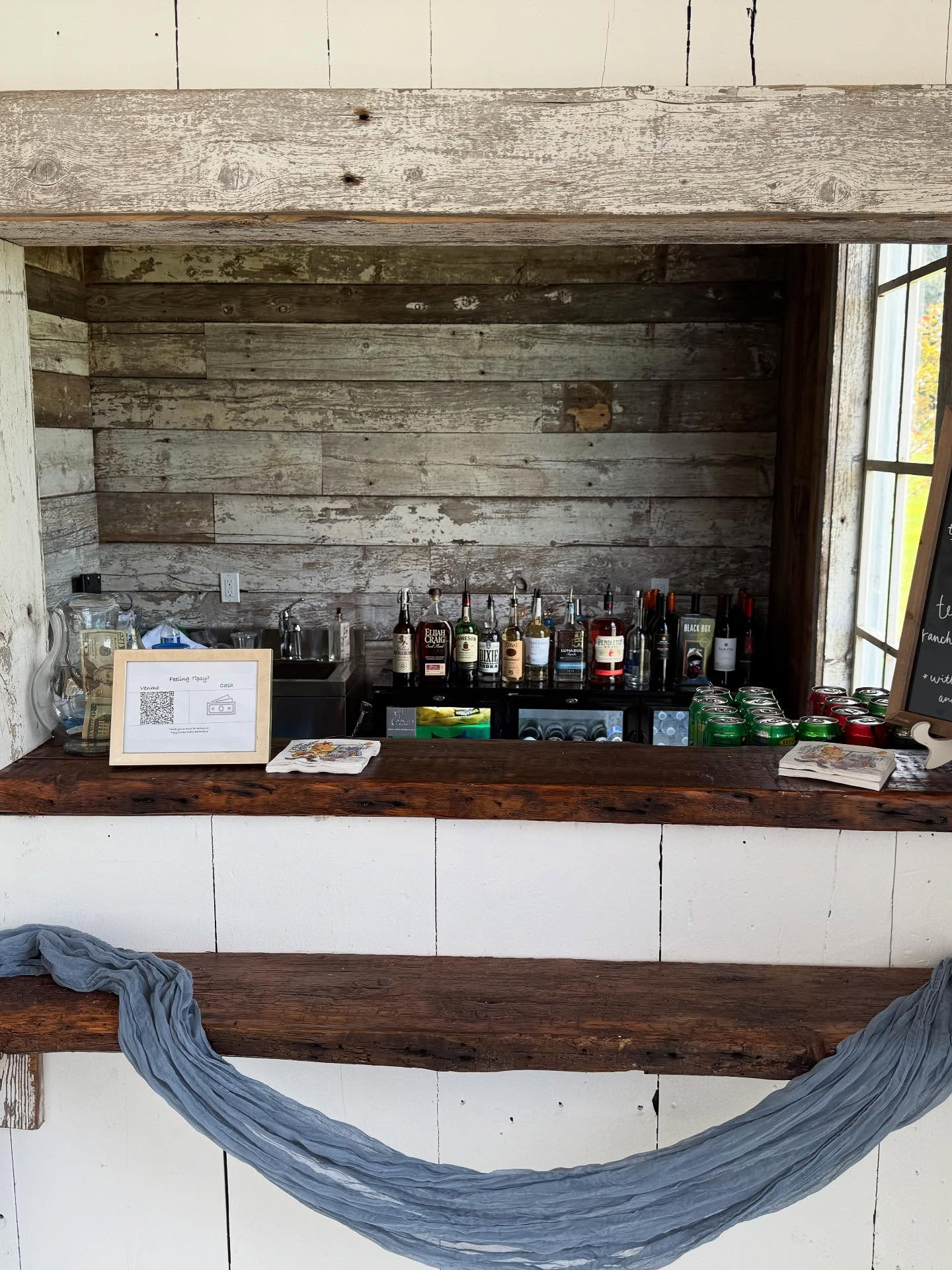 Bar area with assorted liquor bottles, canned drinks, and a blackboard menu, set against a rustic wooden wall.
