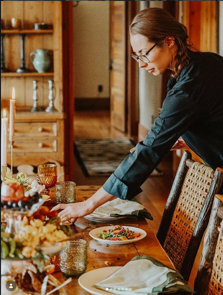 A woman with glasses and braids in a black shirt is setting a dining table with plates, glasses, and candles in a cozy, wood-paneled room.