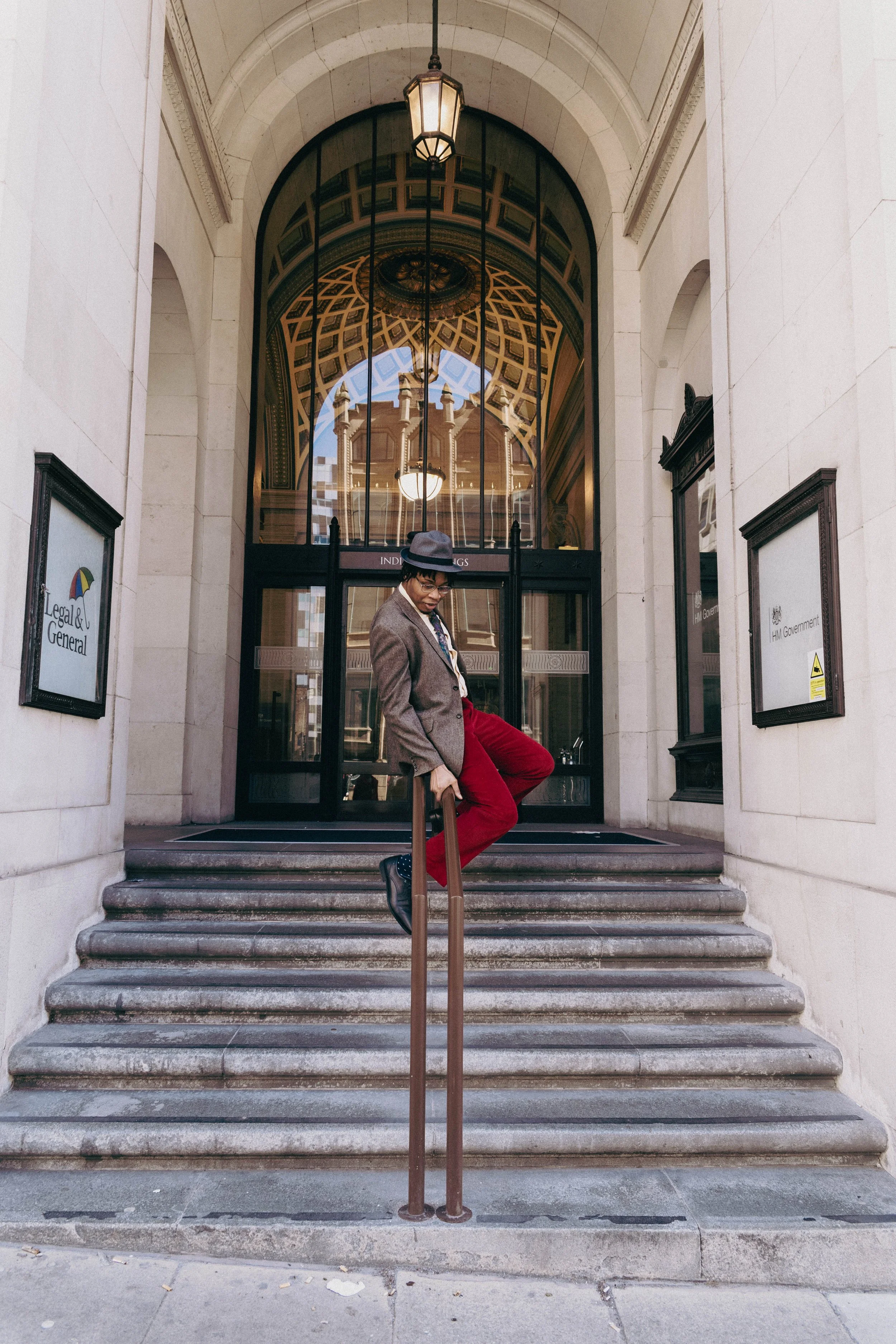 A person dressed in a brown blazer, red pants, and a hat, standing on stairs in front of a large building with ornate archways, glass doors, and decorative ceiling reflections, carrying a suitcase.