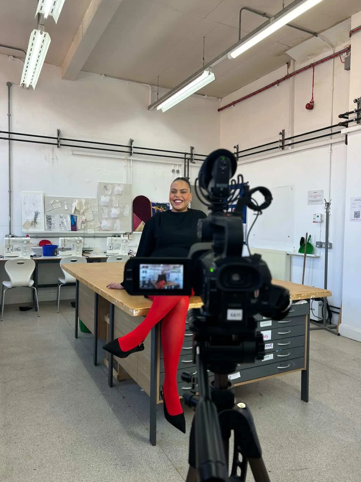 A woman sitting on a desk in a classroom, being filmed by a camera on a tripod. She is smiling and wearing a black top, red tights, and black shoes with red soles. The classroom has white walls, ceiling lights, and various classroom supplies in the background.