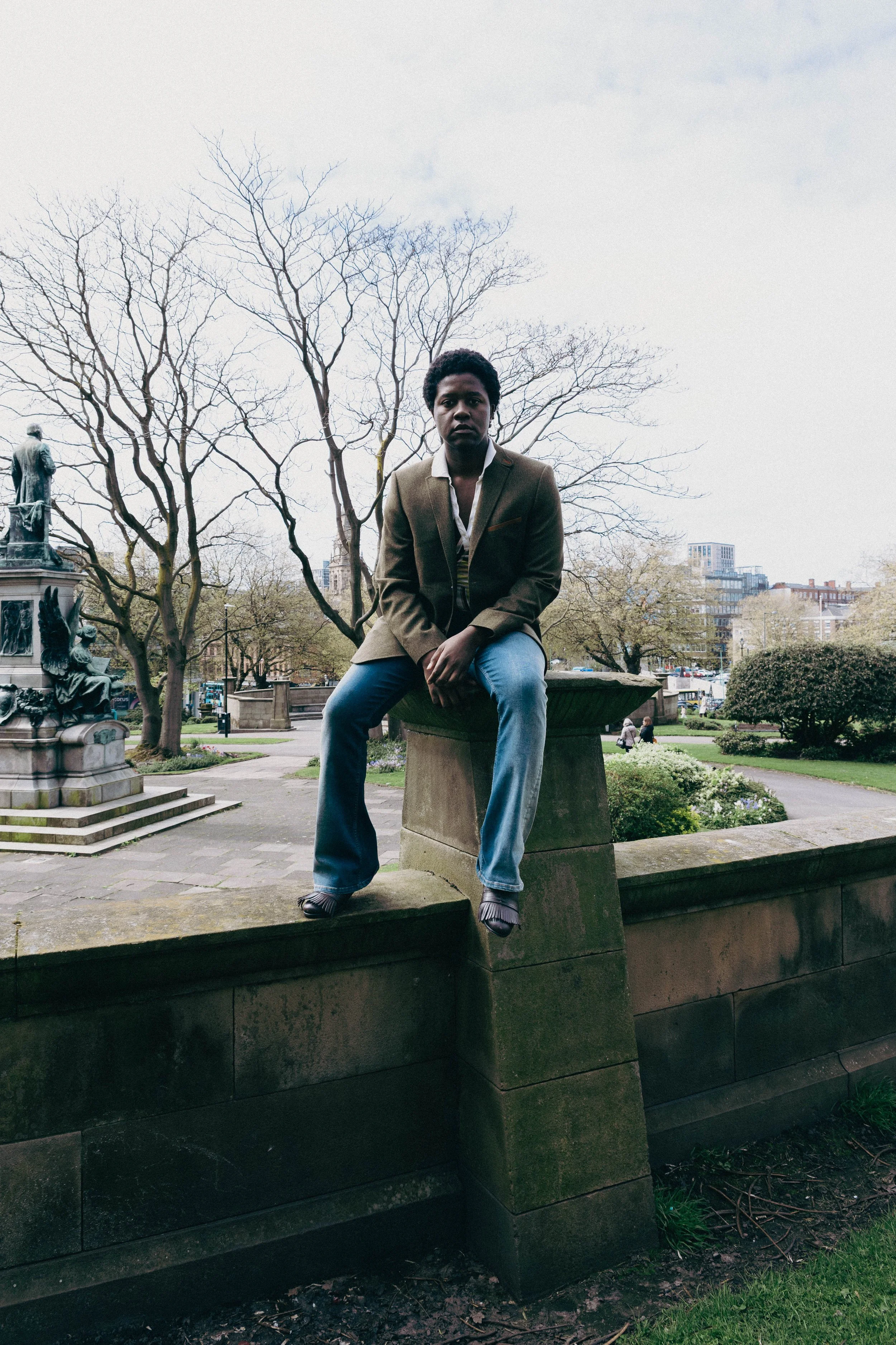 A young man with dark curly hair and medium skin tone sitting on a stone ledge in a park. He wears a brown blazer, white shirt, and blue jeans. Behind him, there are leafless trees and a cloudy sky. In the background, there are buildings and a bronze
