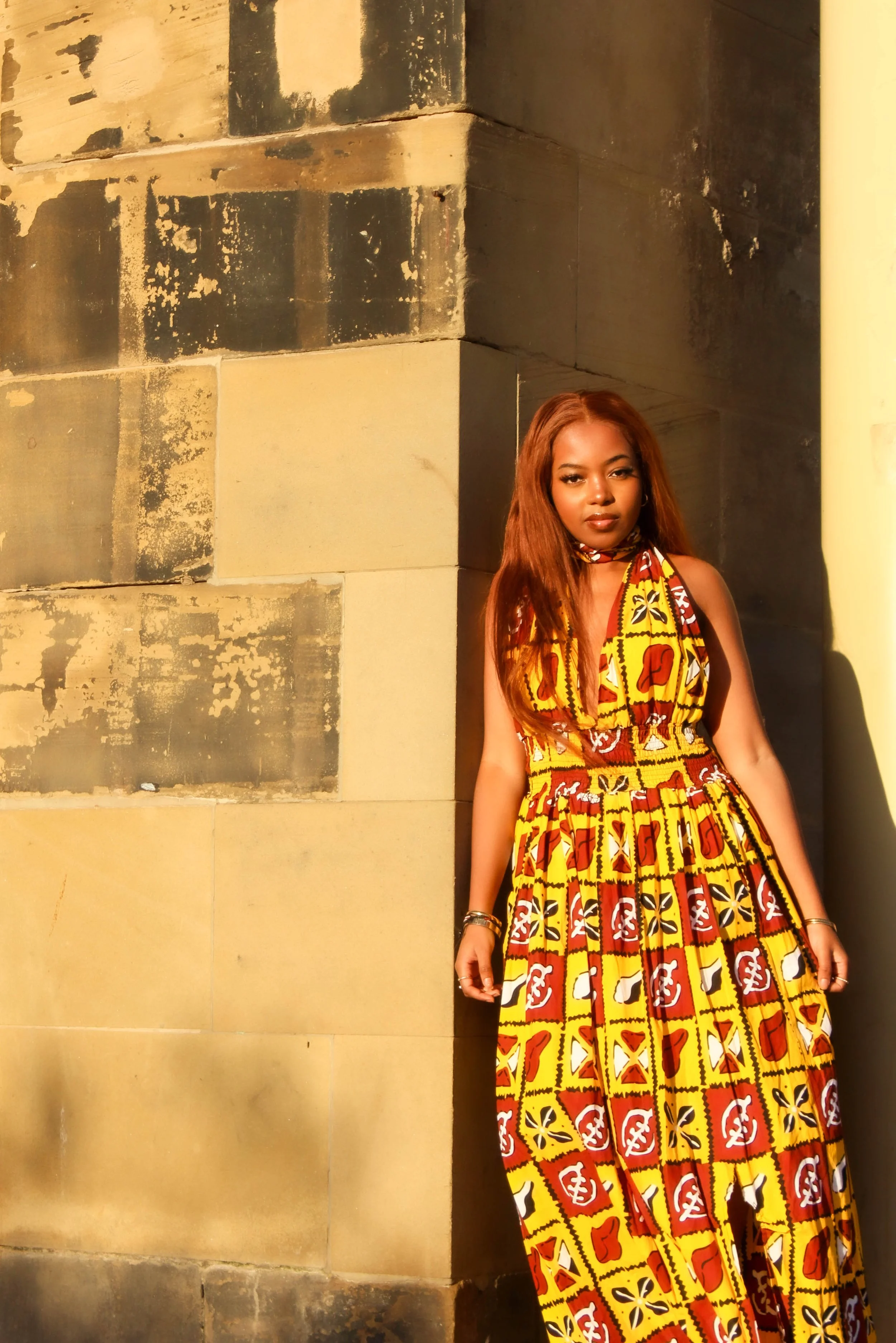 A woman with long reddish-brown hair wearing a colorful African print dress stands against a weathered stone wall at sunset.
