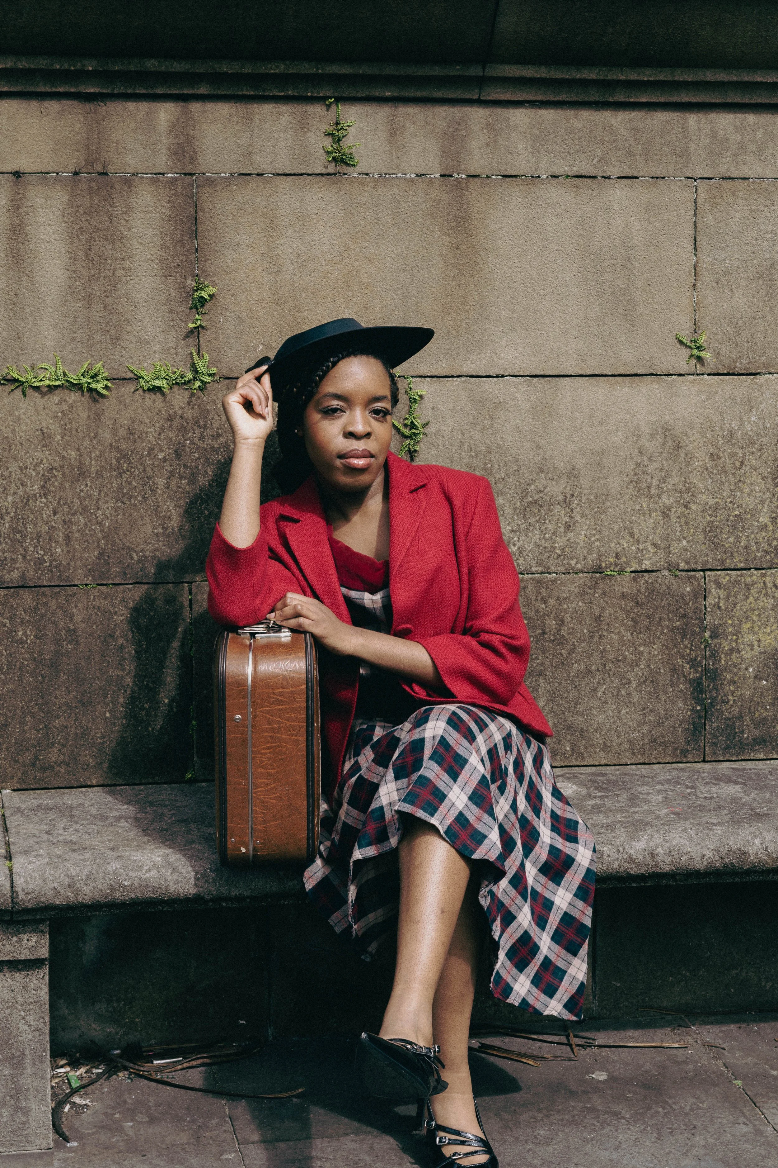 A woman sitting on a stone bench against a brick wall with small green plants growing in the mortar joints. She wears a red blazer, plaid skirt, black heels, and a wide-brimmed black hat, holding a guitar case.