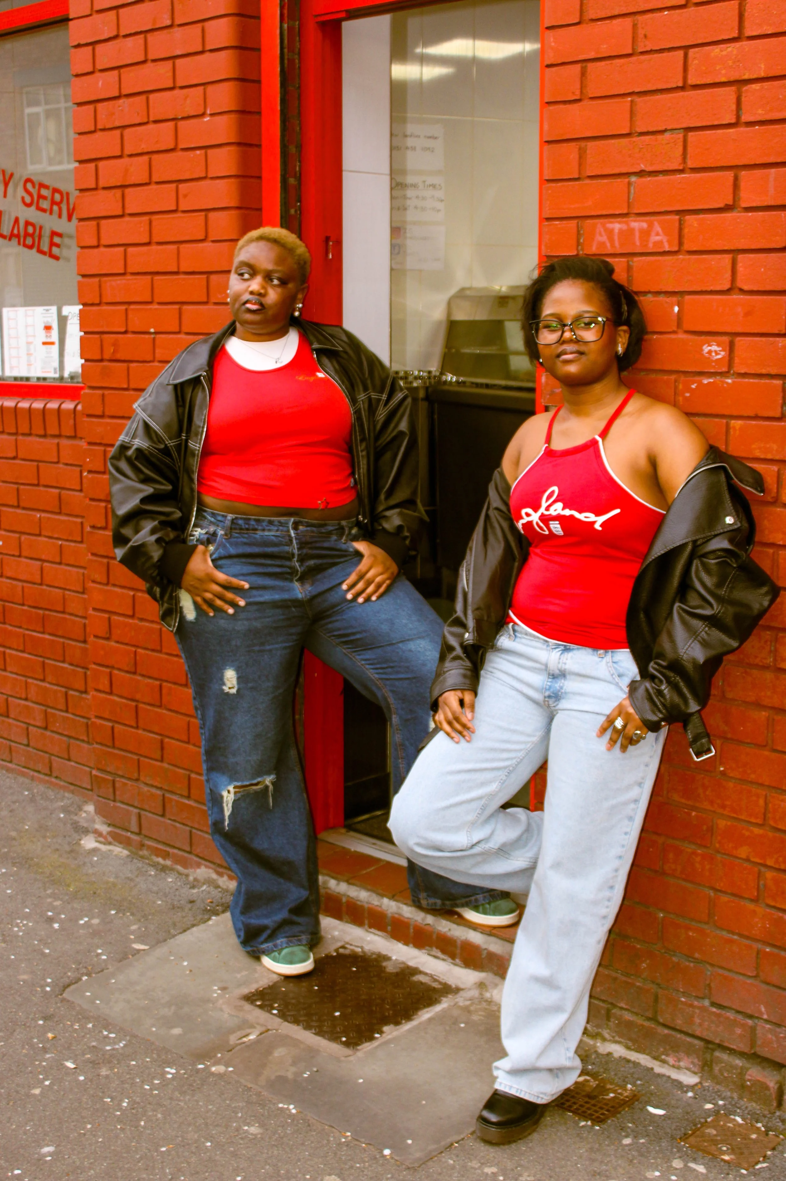 Two women leaning against a red brick wall outside a building. Both are dressed in casual attire with red tank tops, black and gray jackets, and jeans. One woman has short blonde hair and the other has shoulder-length black hair and glasses.