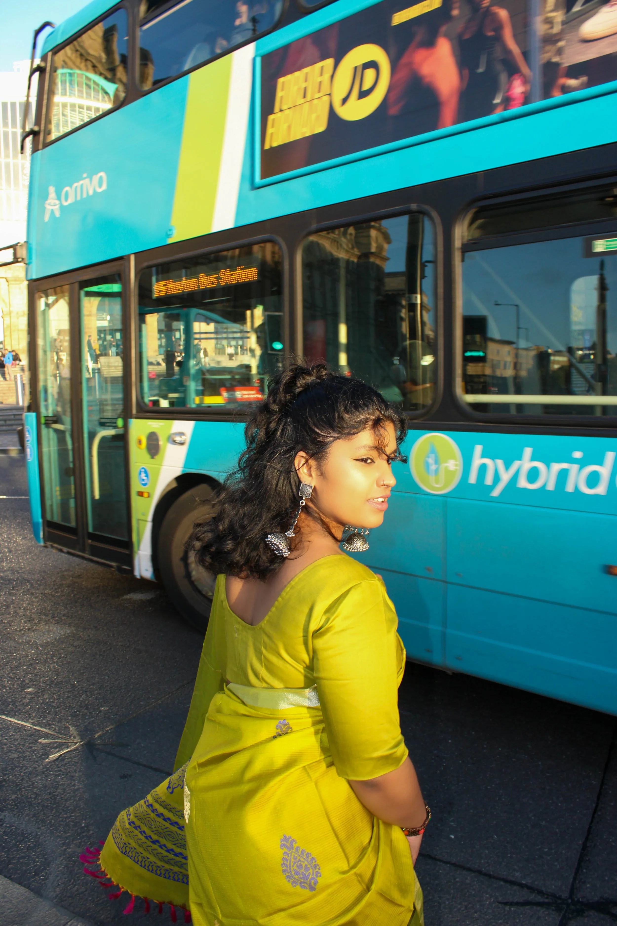 A woman dressed in traditional yellow attire with silver earrings and jewelry, standing on a city street near a blue sightseeing double-decker bus with advertisements and digital displays, with buildings and reflections in the windows.