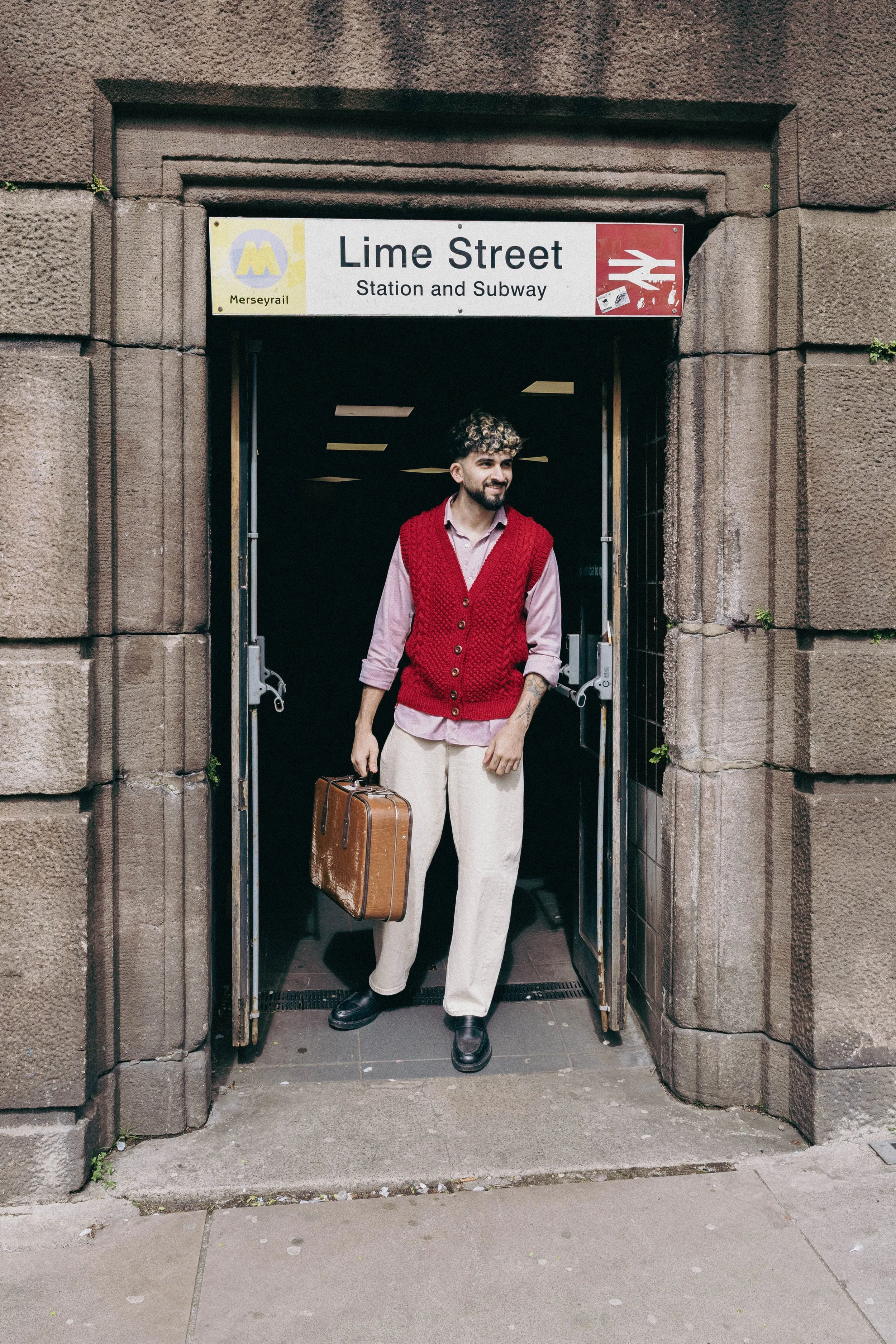 A man in beige pants, black shoes, pink shirt, and red vest holding a brown suitcase, walking out of a subway station entrance labeled Lime Street in Merseyrail, Liverpool, UK.