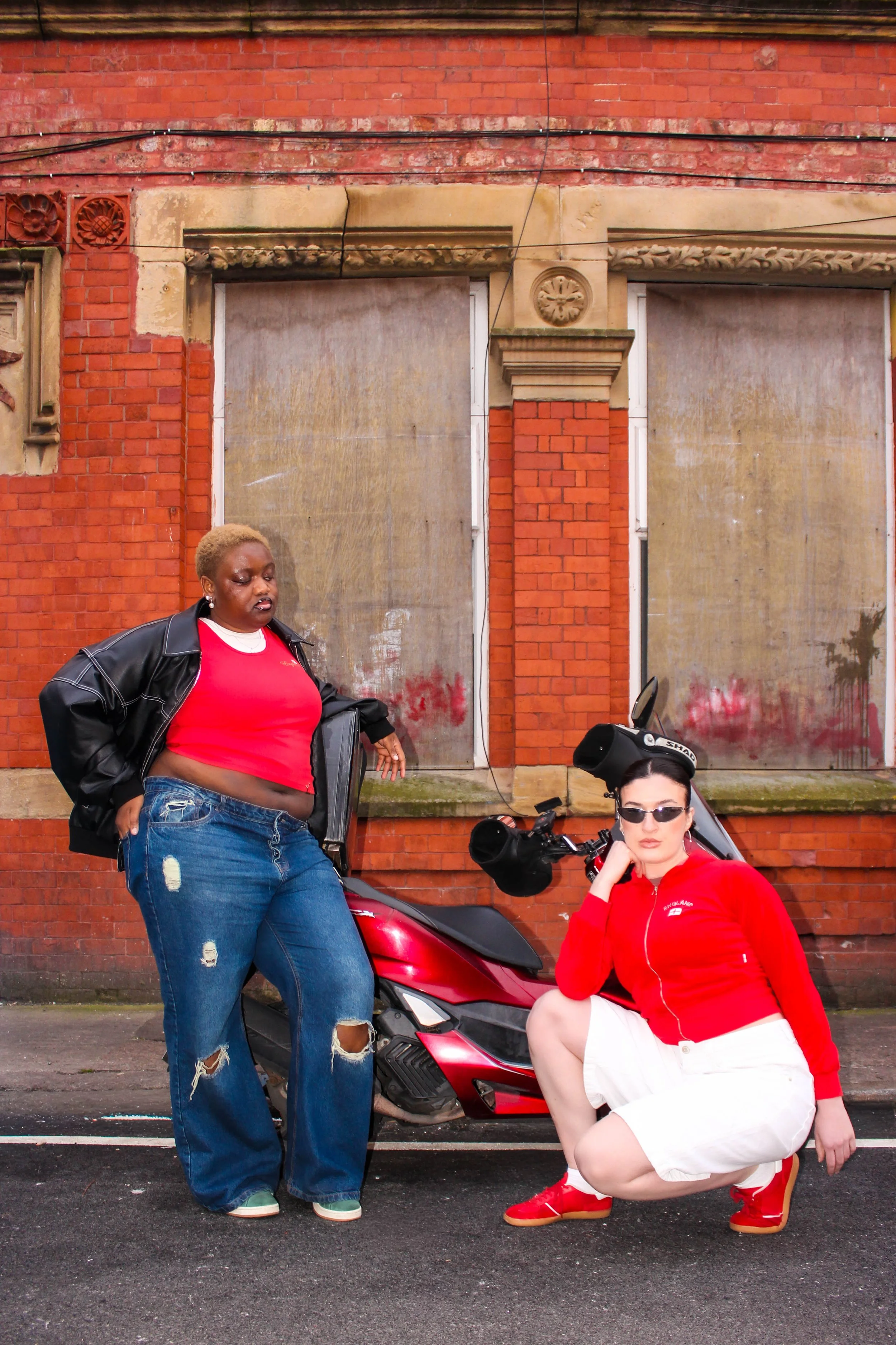 Two women with a red motor scooter in front of a brick and plaster building. One woman stands, the other sits, both wearing red tops. The standing woman has short blonde hair and distressed jeans, the sitting woman has dark hair and sunglasses.
