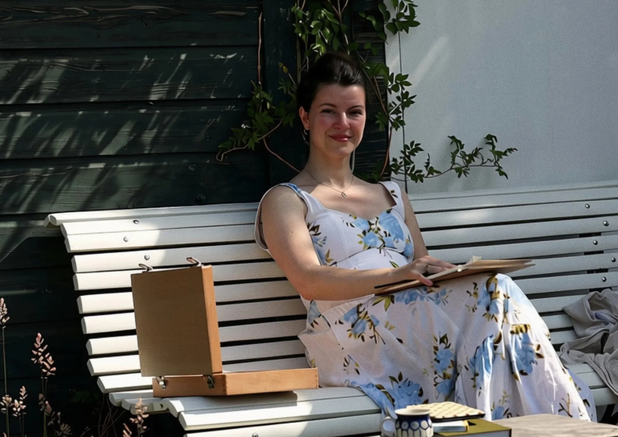 A woman with dark hair and a floral dress sitting on a white bench outdoors, smiling at the camera. There are green plants and shadows in the background.