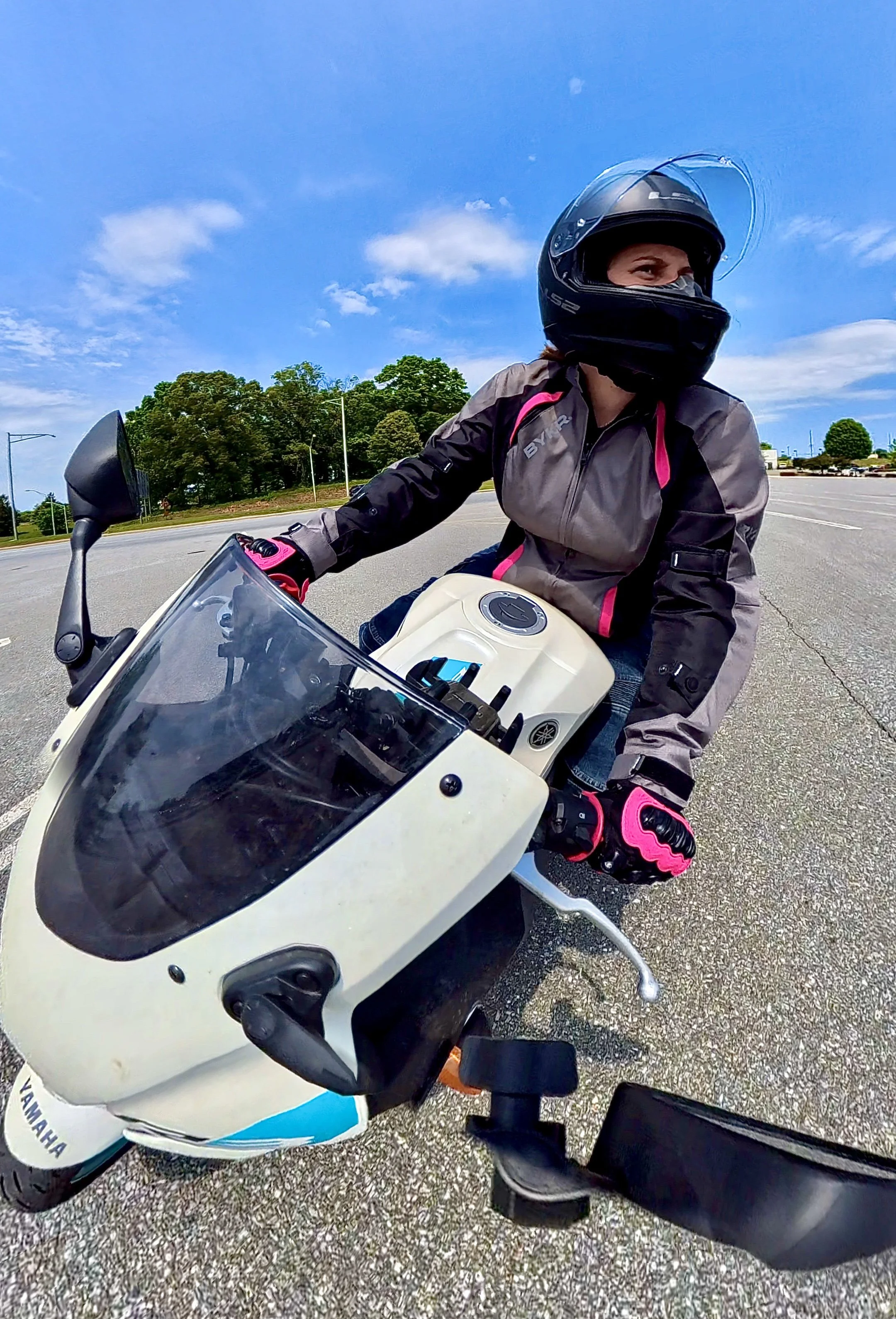 A woman wearing a motorcycle helmet and protective gear riding a Yamaha R3 motorcycle in an open parking lot on a sunny day with a blue sky and scattered clouds.
