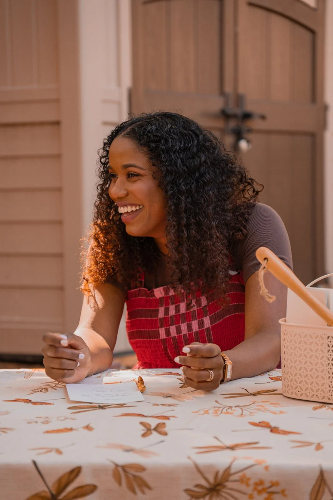 A woman with curly black hair, smiling, sitting at a table with a butterfly-patterned tablecloth, wearing a red and black apron over a brown shirt, with a rolling pin in a basket nearby.