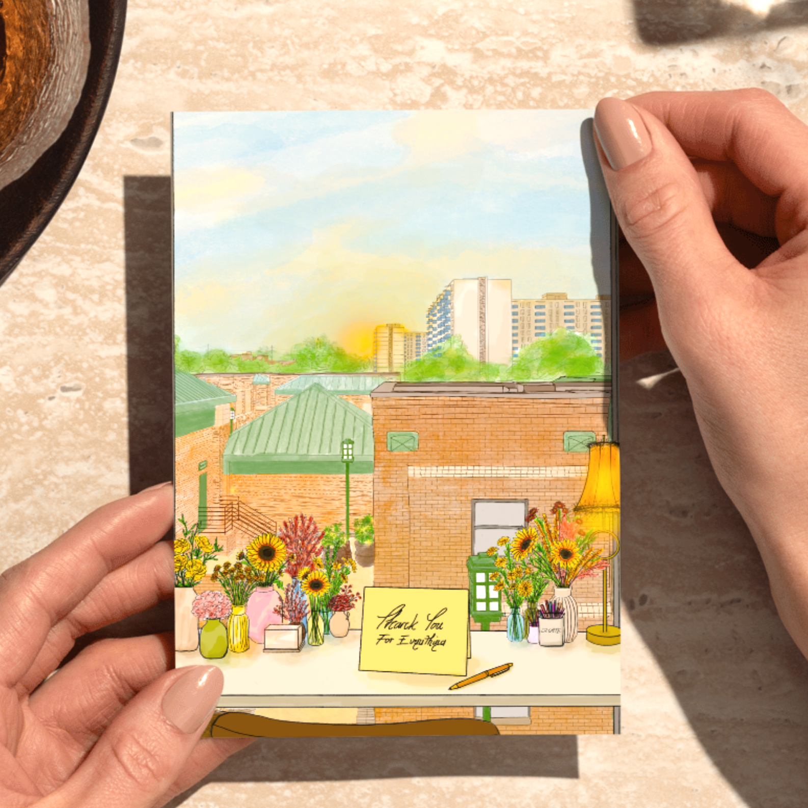 A person holding a colorful illustrated card with a cityscape, flowers, and a 'Thank You for Everything' note on a table with a granite surface.