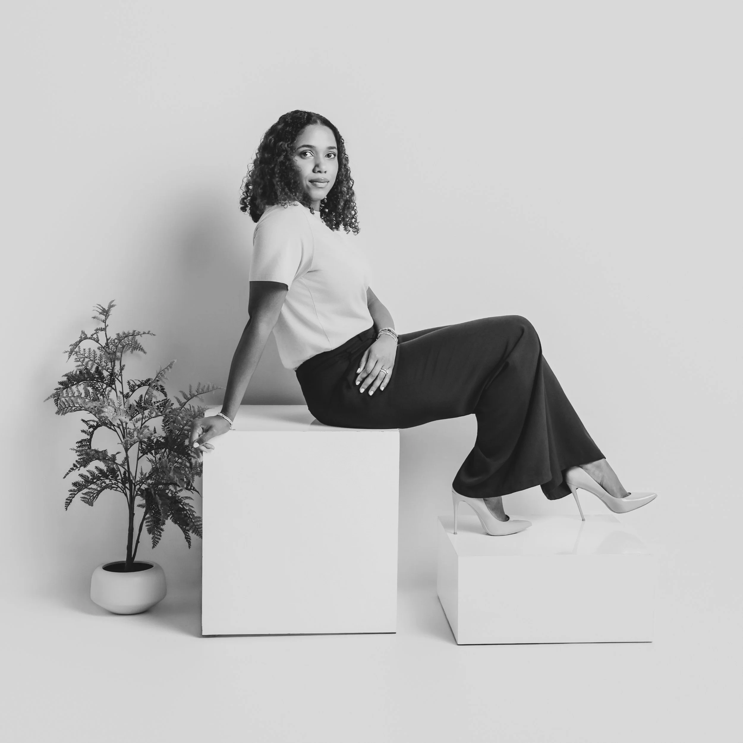 Black and white portrait of a woman sitting on a white block with a potted plant nearby, against a plain light gray background.