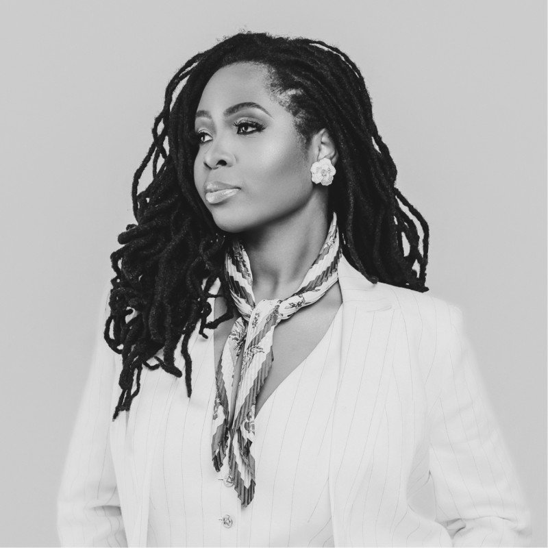 Black and white portrait of a woman with long dreadlocks, wearing a striped scarf, floral earrings, and a pinstripe blazer.