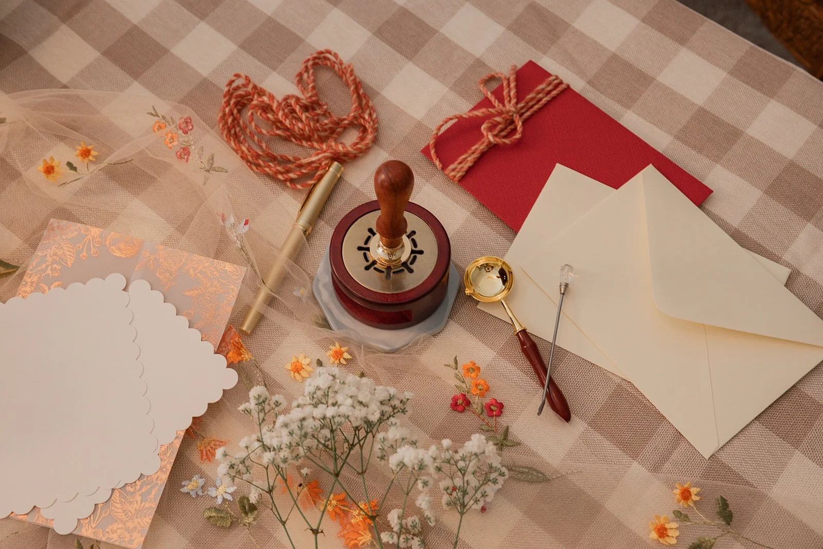 Decorative items for a celebration or event on a checkered tablecloth, including a wooden rubber stamp, a small gold ink pad, a red envelope tied with string, a white envelope, flower cutouts, and small artificial flowers.
