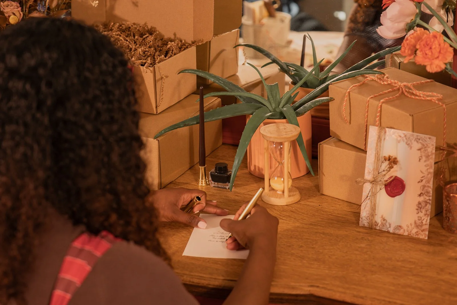A person with curly hair writing a note at a table decorated with potted plants, gift boxes, a small hourglass, a candle, a pen, and a greeting card with a red wax seal.