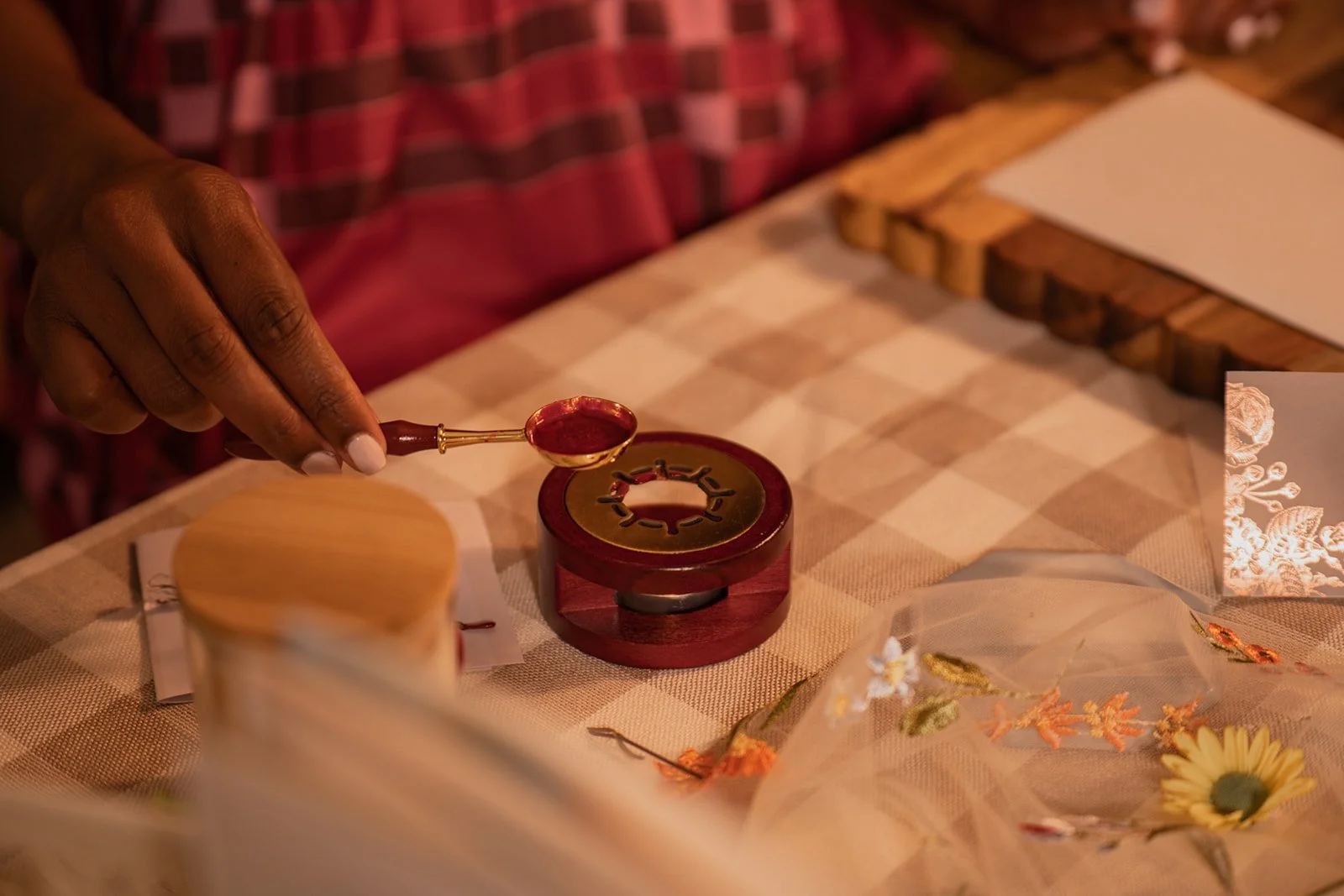 A person uses a small stick to pour wax into a circular wax melting device at a table with a checkered tablecloth, surrounded by decorative flowers and paper items.