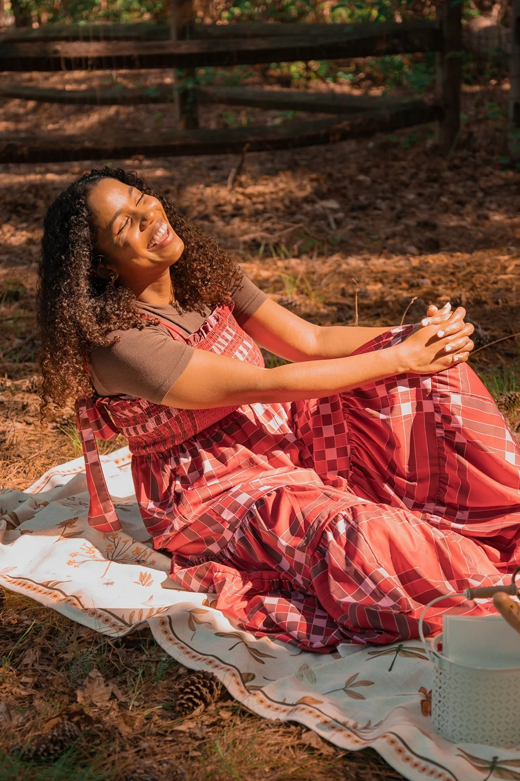 A woman with curly hair sitting on a blanket in a wooded area, smiling with her eyes closed as she holds her hands together.