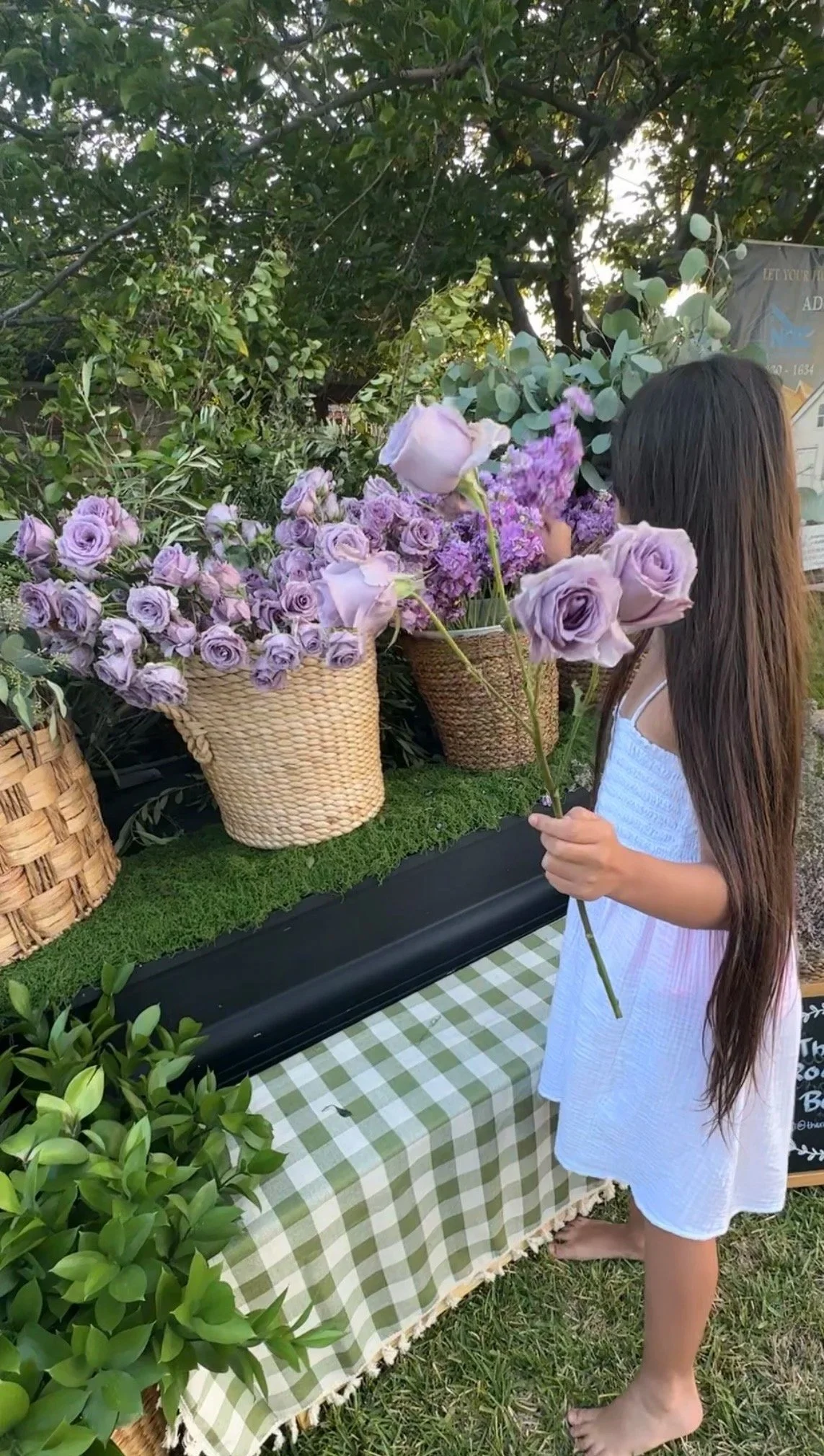 A young girl with long brown hair in a white dress holding a bouquet of light purple roses at an outdoor flower stand with baskets of flowers and greenery.