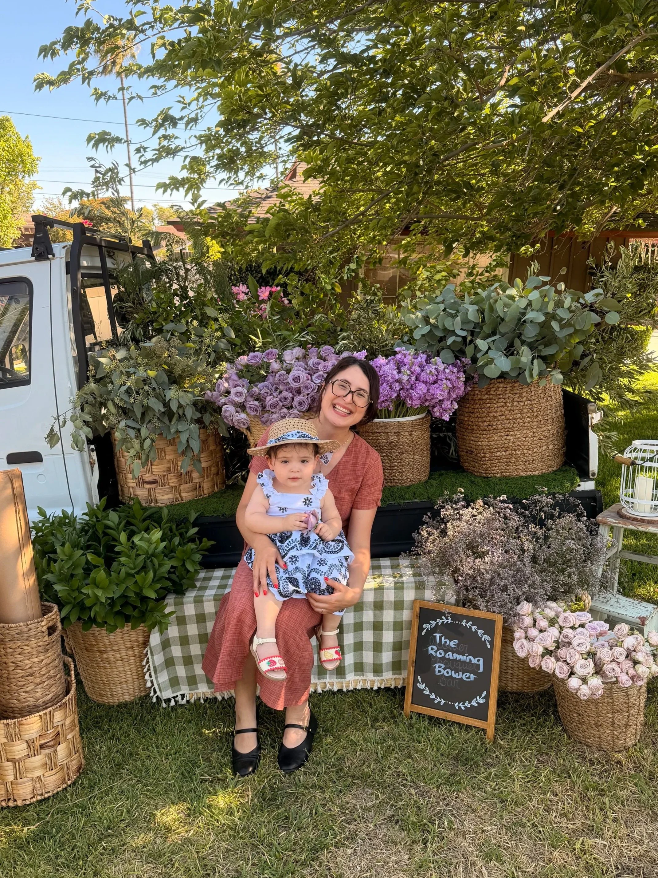 A smiling woman in a brown dress holding a young girl in a white dress with blue patterns, both sitting in front of a flower bouquet display at an outdoor event with green grass and trees. There is a chalkboard sign reading 'The Roaming Roasting Bowery'.