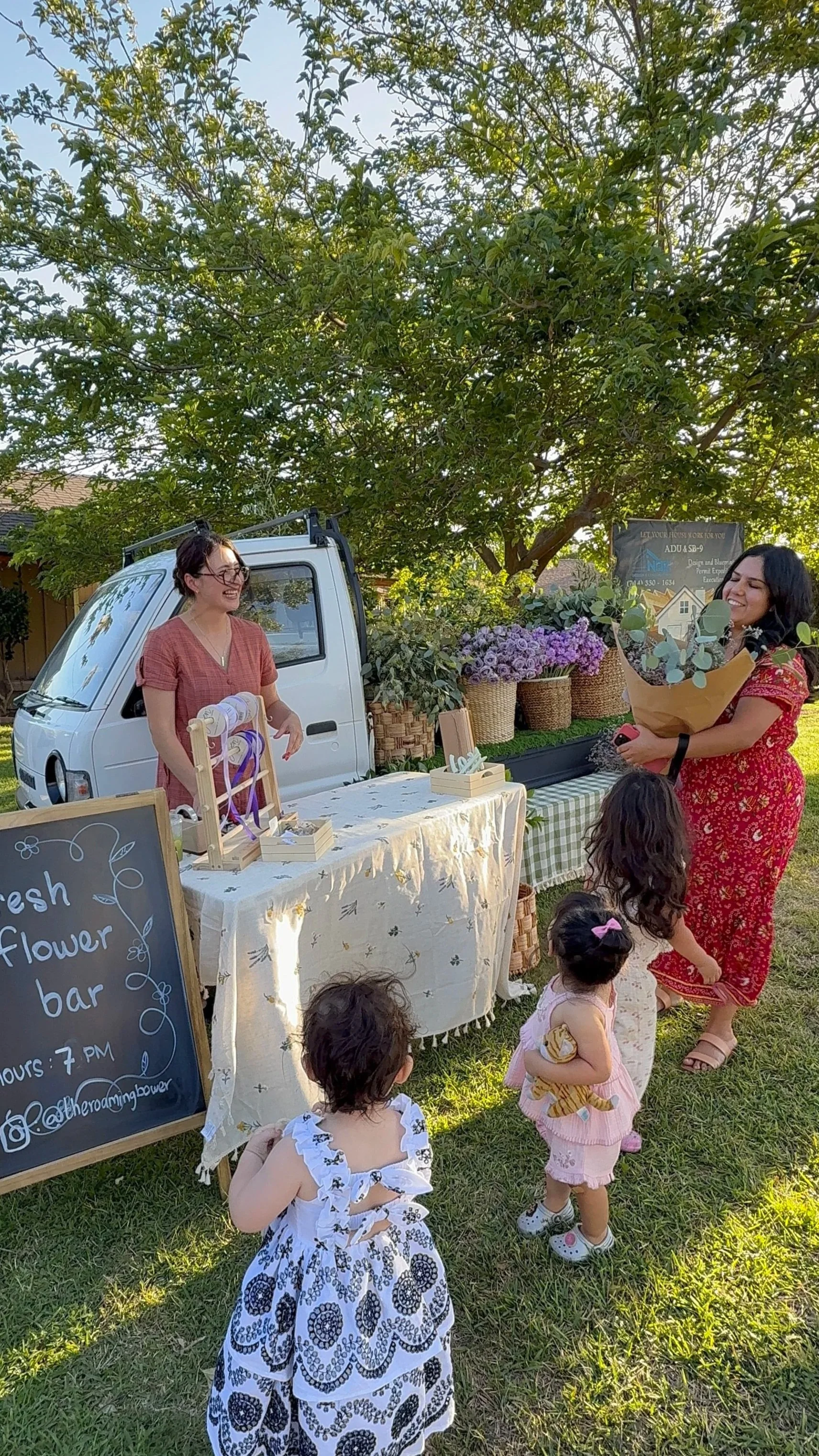 A woman with short dark hair and glasses, wearing a red dress, is standing behind a table at an outdoor flower bar, smiling at a woman with long dark hair in a red dress holding a bouquet of flowers. Several children are in the foreground, looking towards the women. The table has flower arrangements, decorations, and a sign that reads "Fresh Flower Bar." Behind, a small white van is parked with large baskets of flowers and greenery, under a large leafy tree on a grassy yard.