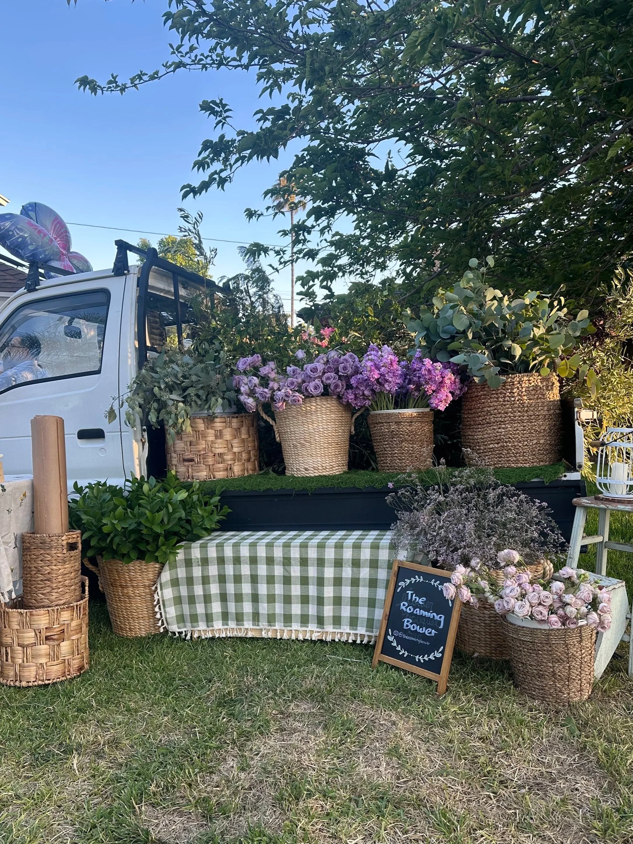 Market stall with woven baskets filled with purple and pink flowers, a chalkboard sign that reads 'The Roaming Flower Bow,' a table with a green and white checkered tablecloth, and various green plants against a backdrop of trees and a clear blue sky.