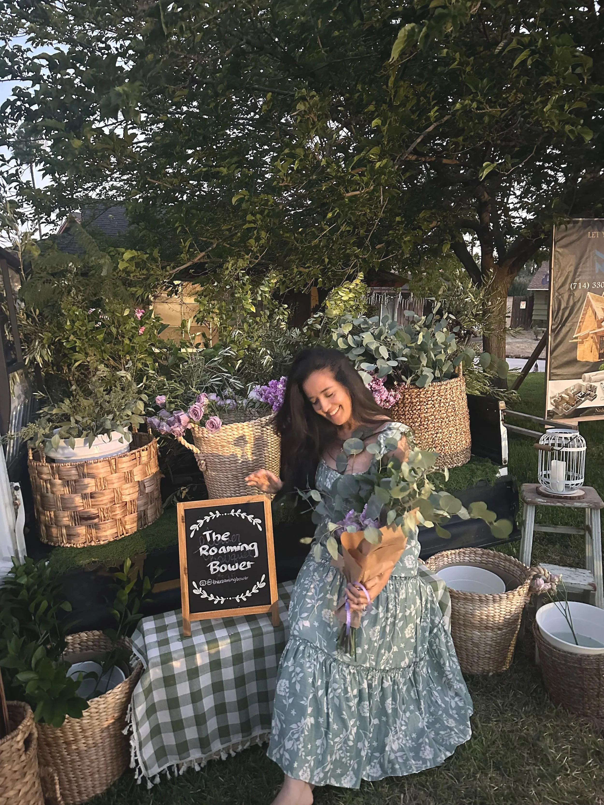 A woman holding a bouquet of flowers at a market stand decorated with green and purple flowers and various baskets, with a sign reading "The Roaming Bower."
