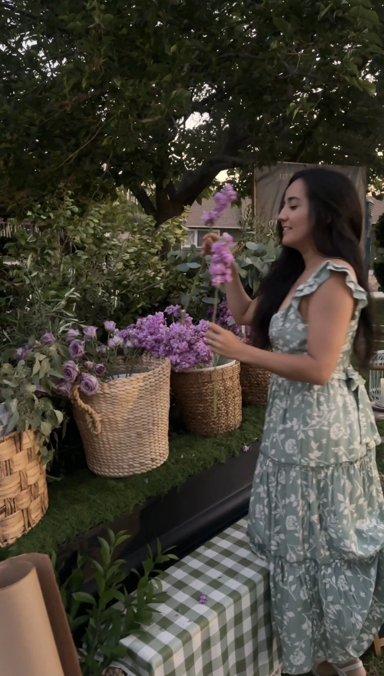 A woman with long dark hair in a blue and white floral dress arranging pink flowers at an outdoor flower stand with baskets of flowers, under a large tree during sunset.