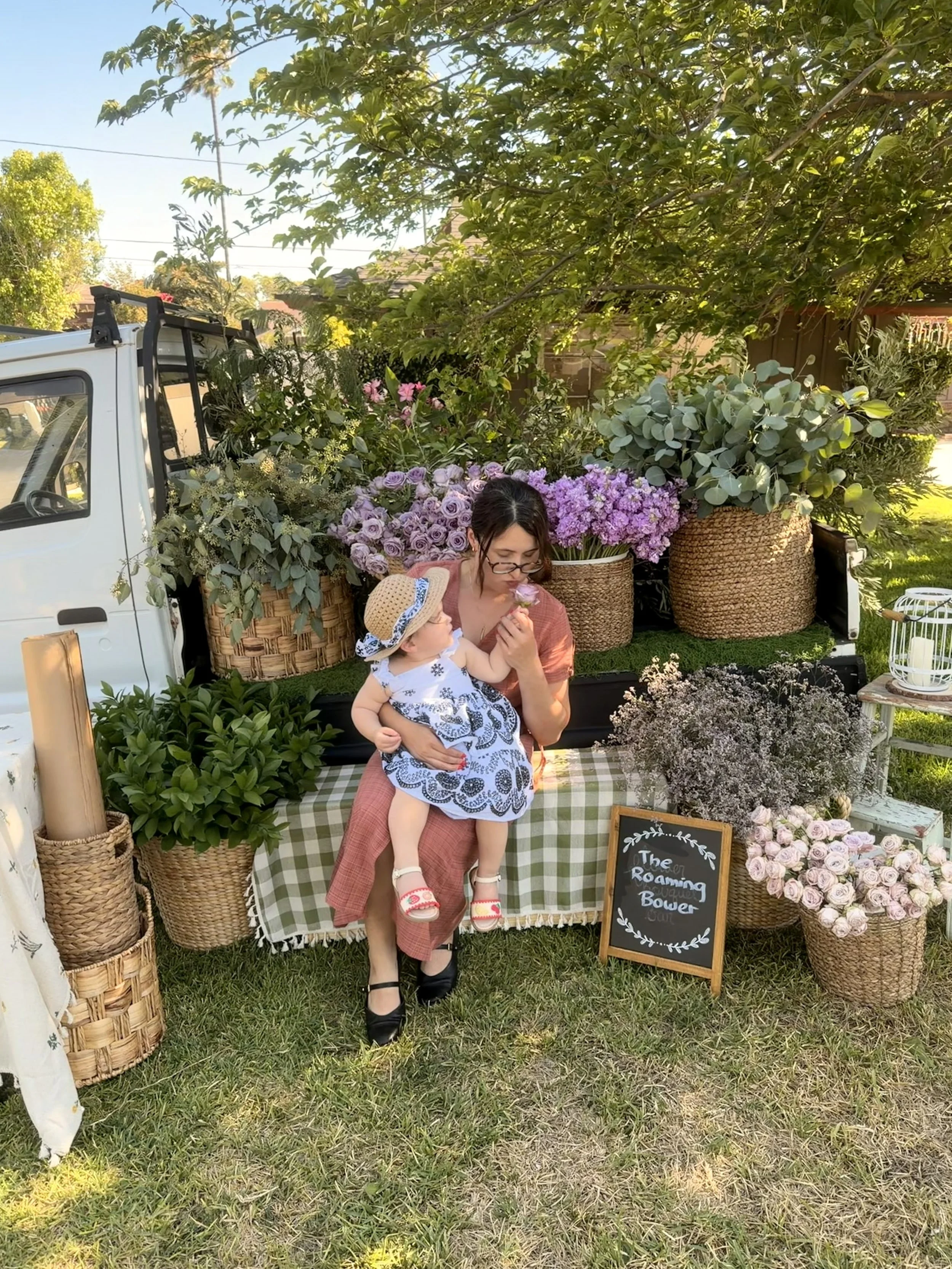 A woman and a child sitting in front of a flower market stand with various plants and flowers, including pink, purple, and white blooms, on a grassy lawn in a sunny outdoor setting.