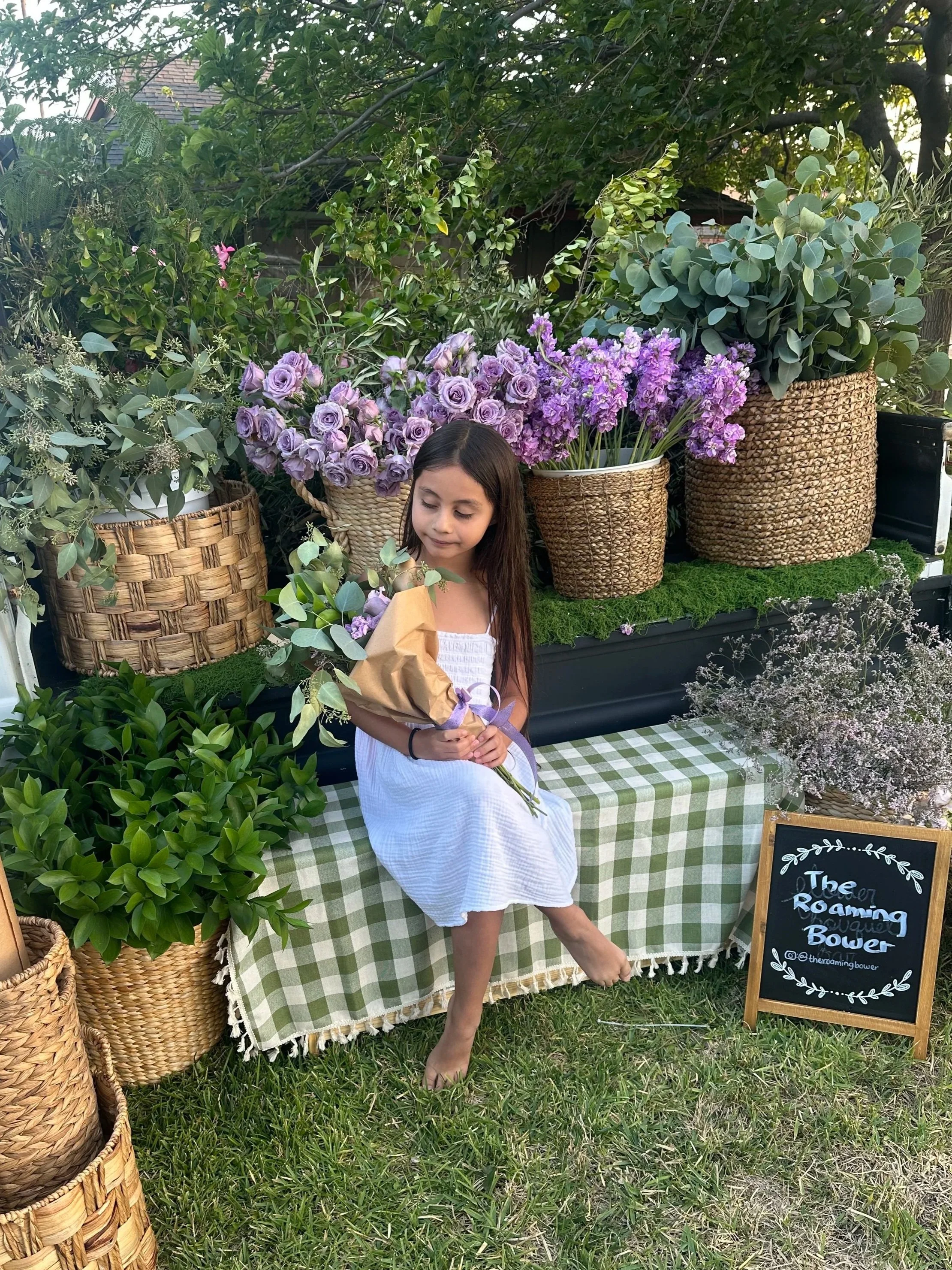 Young girl with long dark hair in a white dress, holding a bouquet of flowers, standing beside a flower display with purple roses and other plants, in an outdoor garden setting.