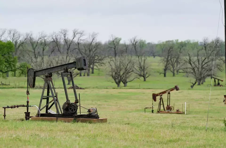 Two sets of oil pumpjacks in a grassy field with bare trees and cloudy sky in the background.