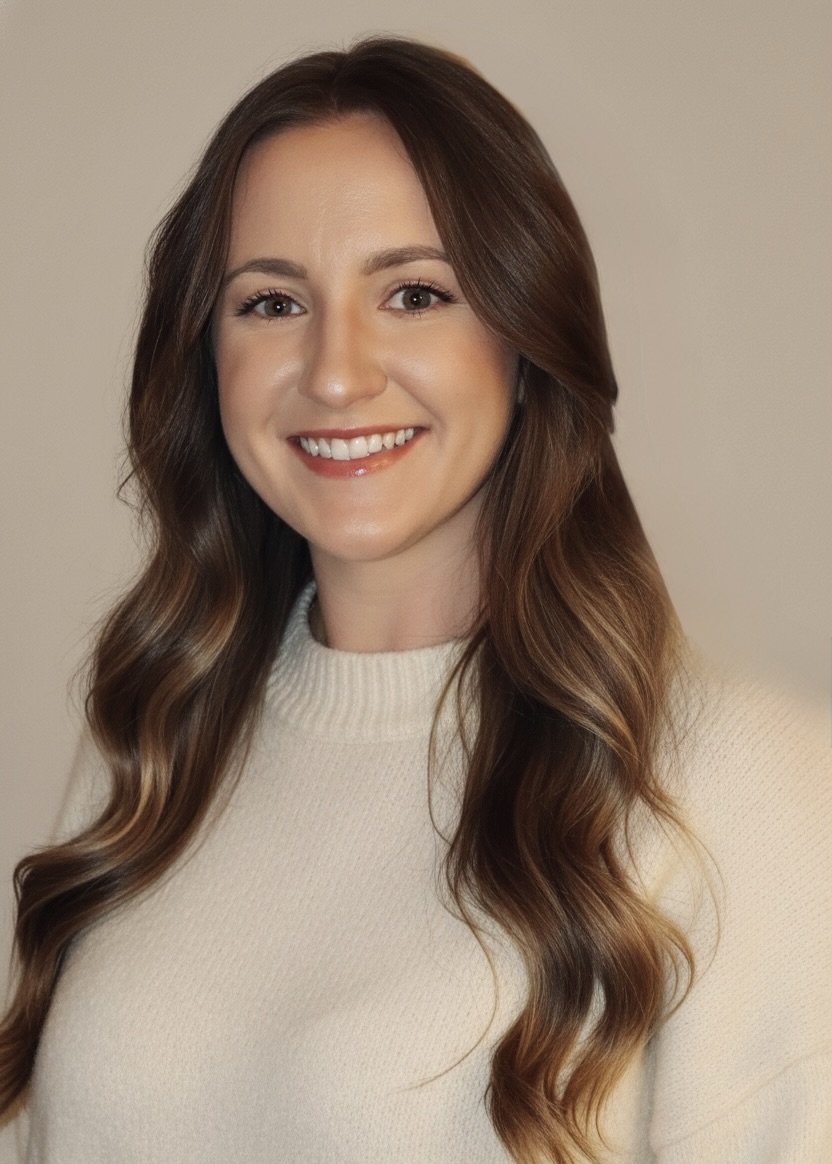 A woman with long, wavy brown hair and a bright smile, wearing a cream-colored sweater, standing against a plain neutral background.