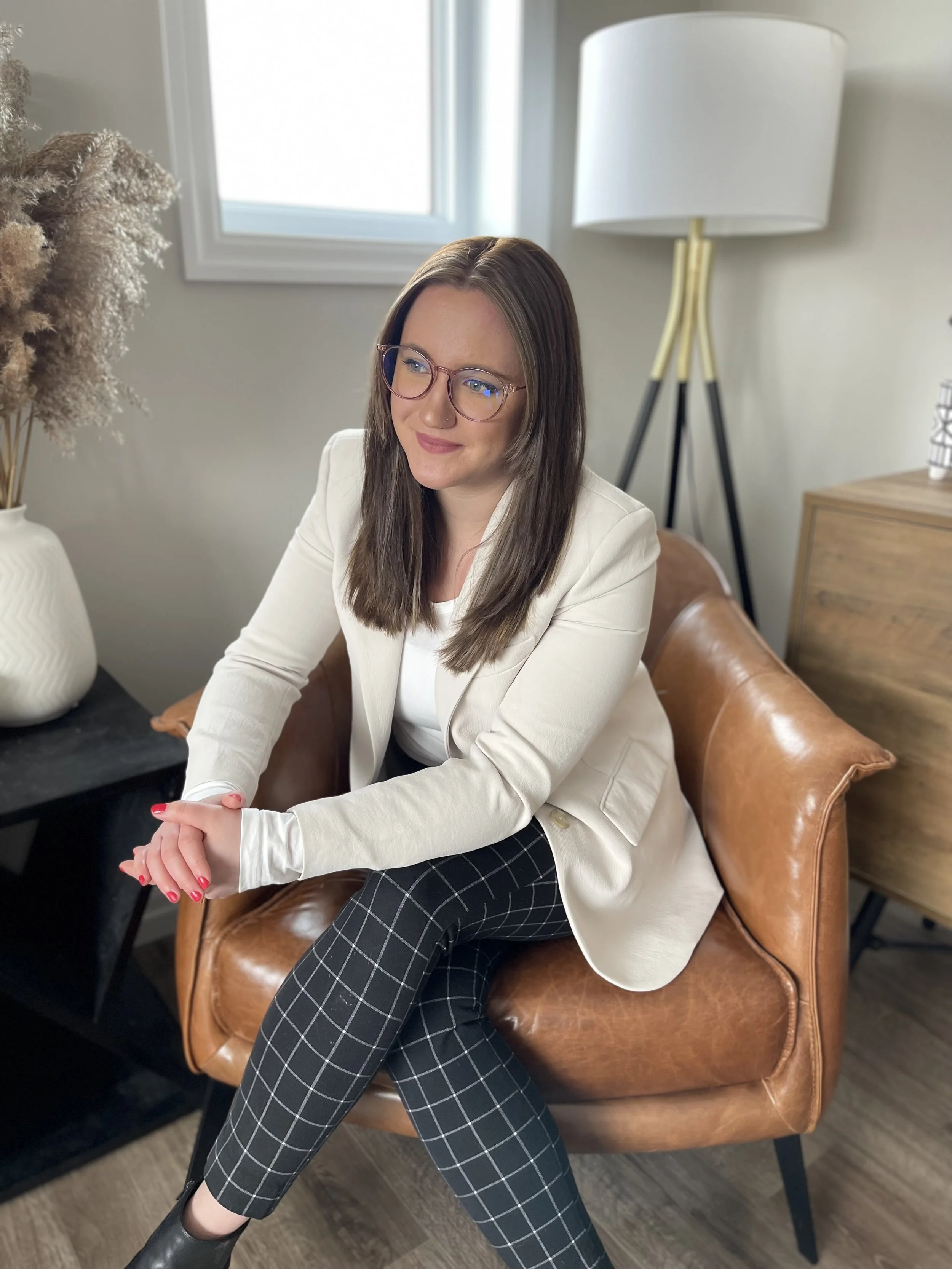 A young woman with brown hair and glasses sits on a brown leather chair, smiling and looking to her right. She is wearing a white blazer, a white top, and black plaid pants. Behind her, there's a large white lamp with a gold and black tripod base, a wooden side table, and a window with gray trim. To her left, there's a white vase with dried pampas grass on a black surface.
