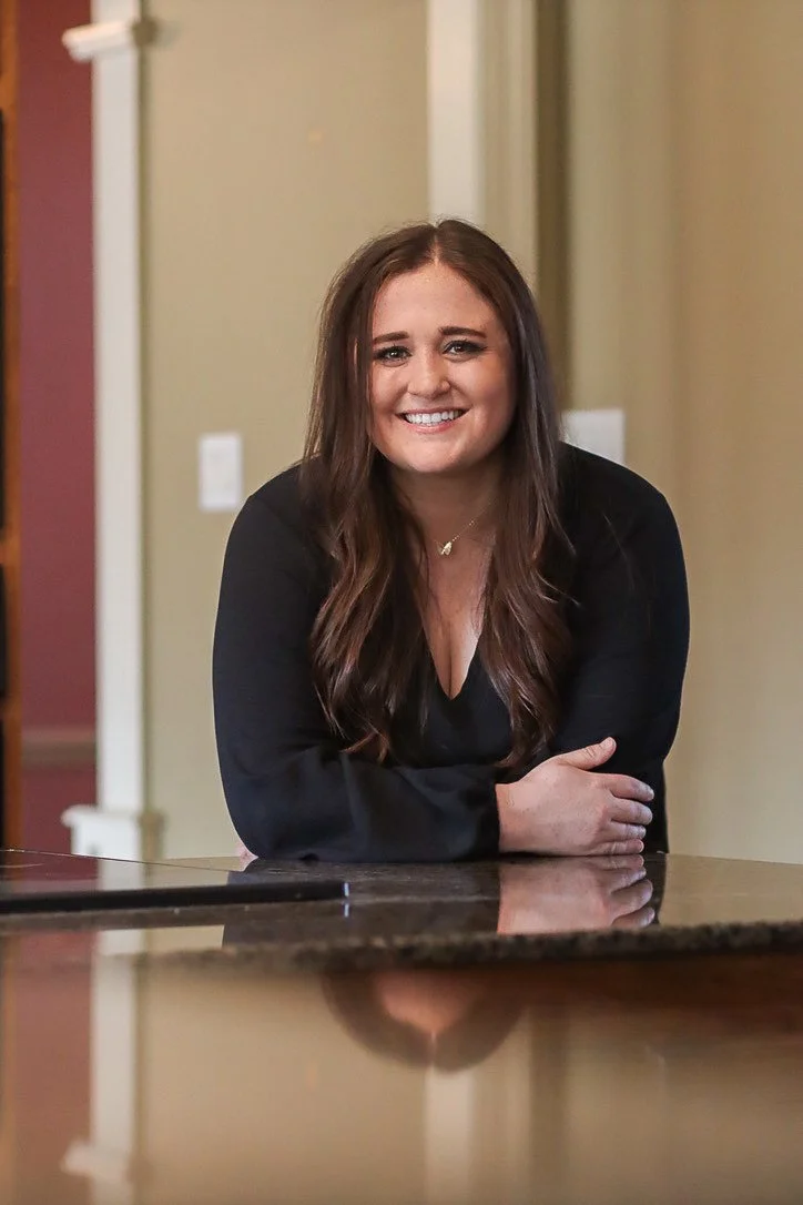 A young woman with long brown hair, smiling, leaning on a kitchen counter, wearing a black top.