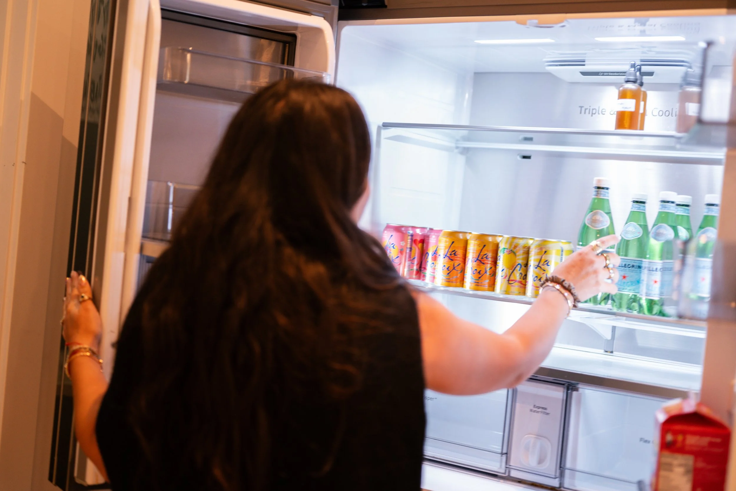 Person with long dark hair looking inside an empty refrigerator with a few bottles and cans on the shelves.