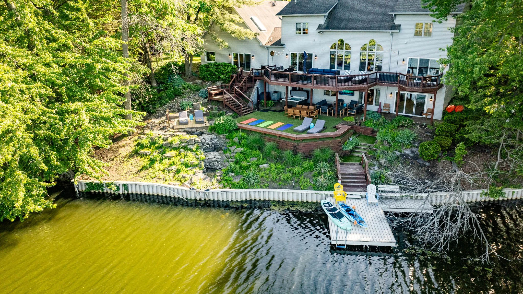 Aerial view of a lakeside backyard with a deck, outdoor furniture, a boat dock, and surrounding lush greenery.