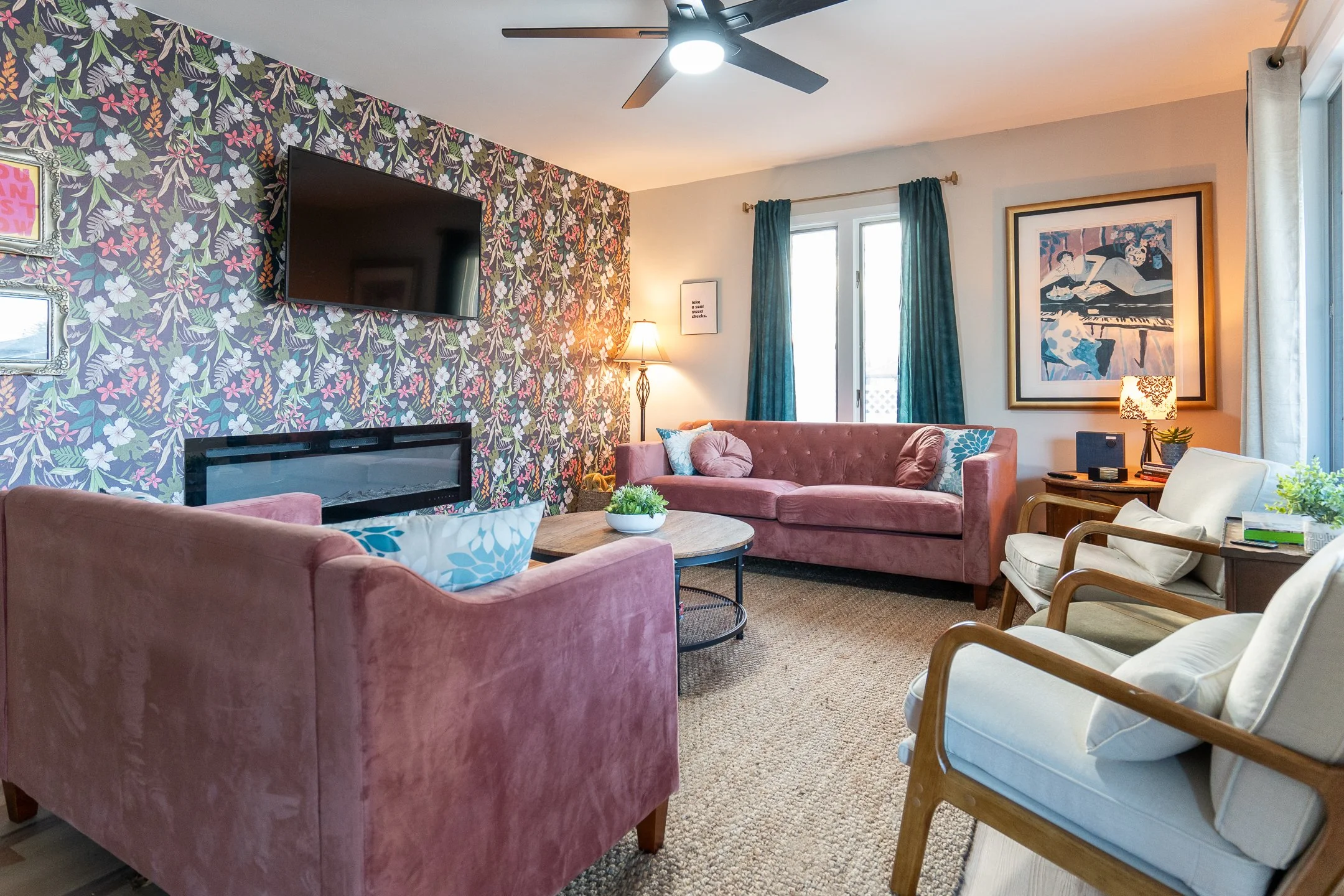 Living room with pink velvet sofa, white armchair with wooden arms, a wall-mounted flat-screen TV, a fireplace, framed art, teal curtains, a standing lamp, and various decor items.