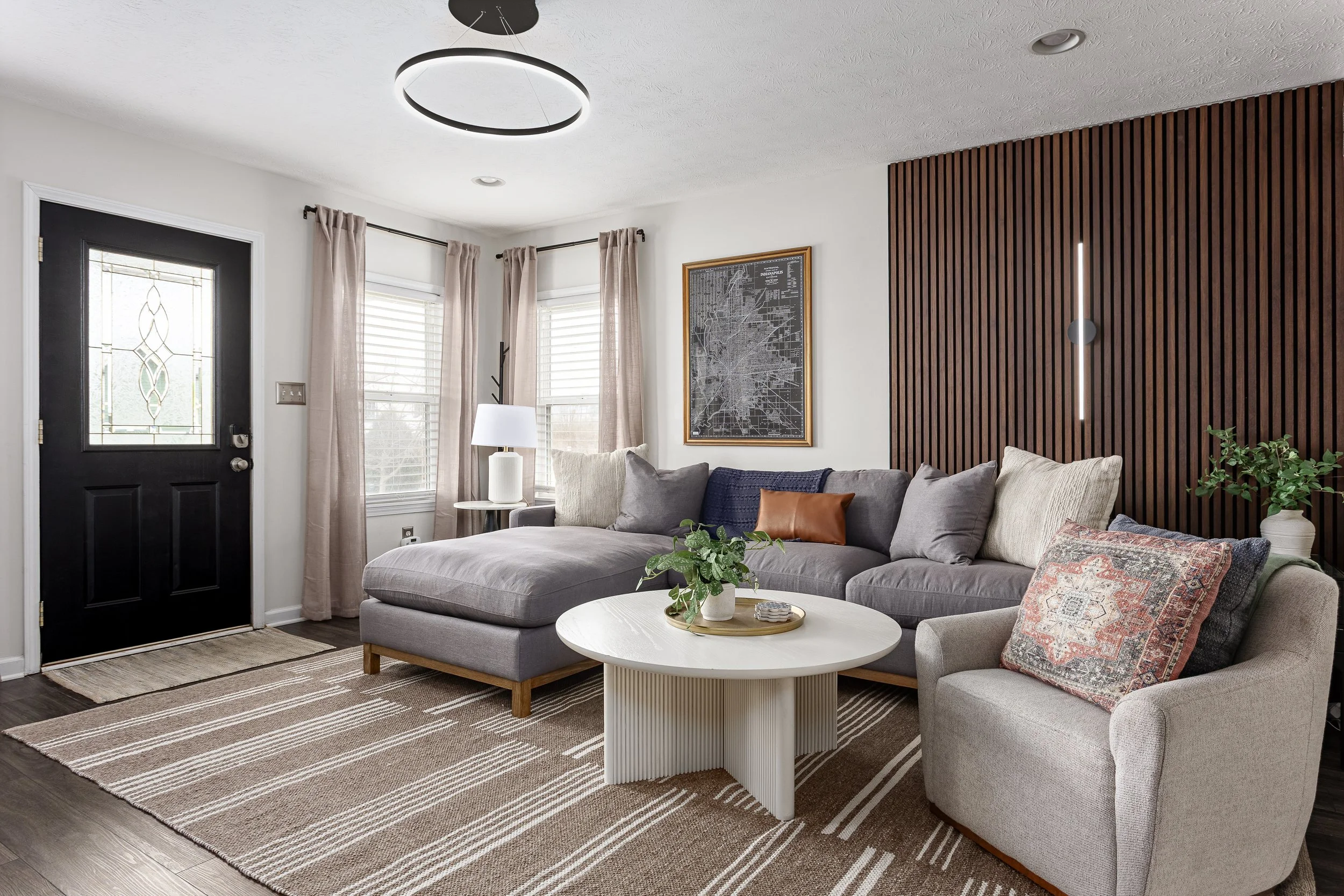 Living room with gray sectional sofa, pillows, white coffee table with plants, beige rug, framed map on wall, beige curtains, black front door with decorative glass, ceiling light, wooden accent wall, window with blinds, lamp, and potted plant.