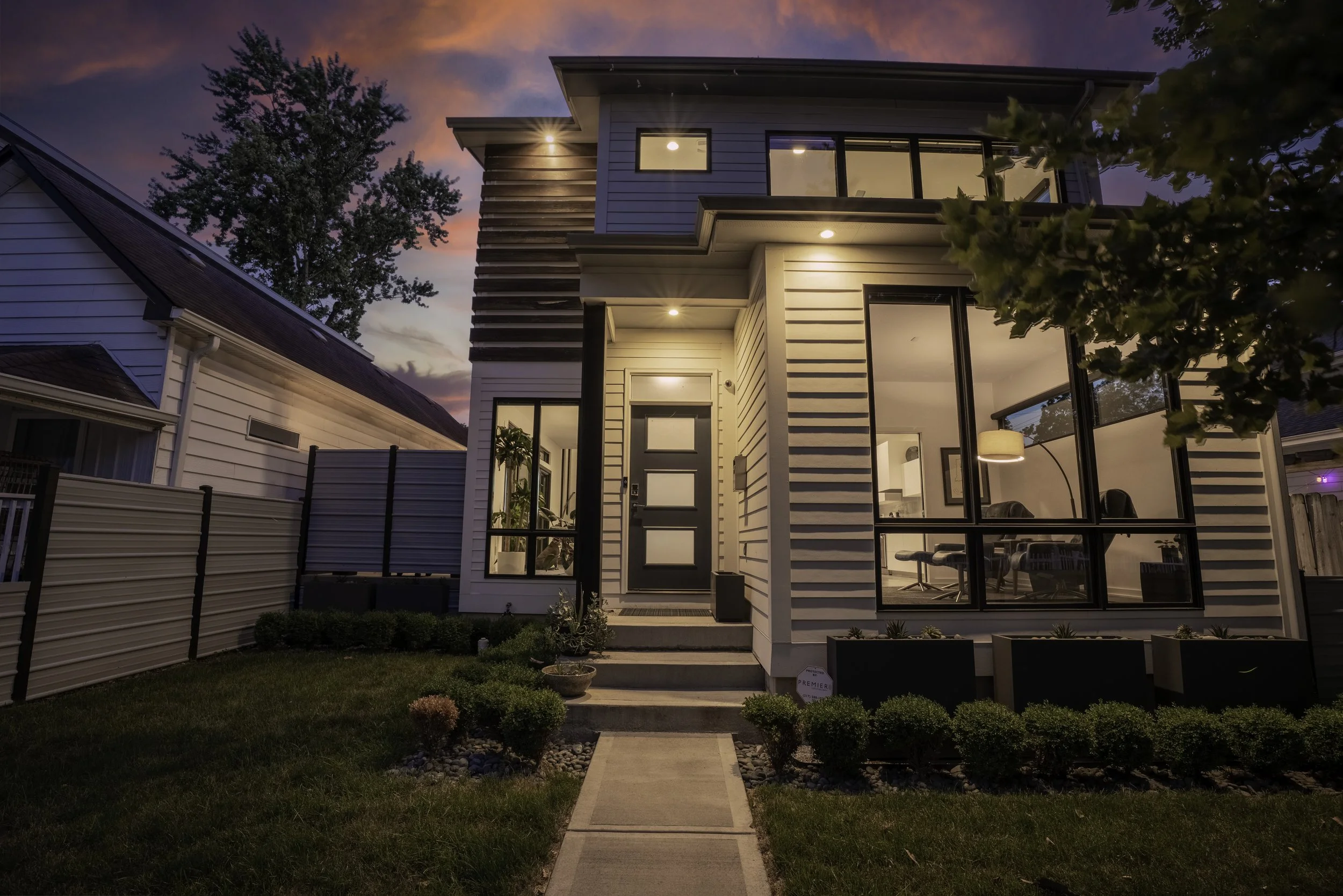 Modern two-story house with large windows, front yard with pathway, and landscaping, during sunset with colorful sky.