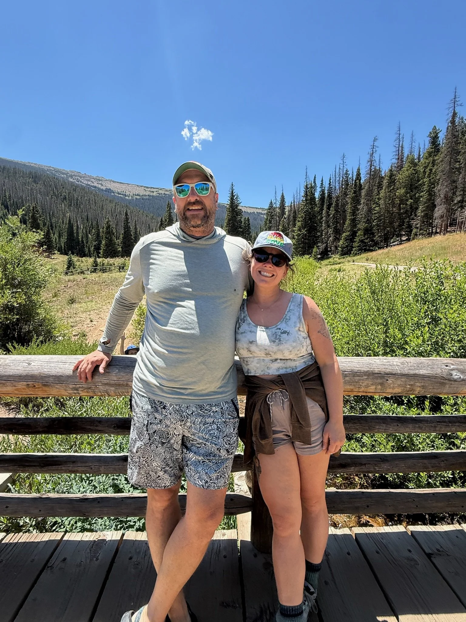 A man and woman posing together outdoors on a wooden platform, with forested mountains and a clear blue sky in the background.