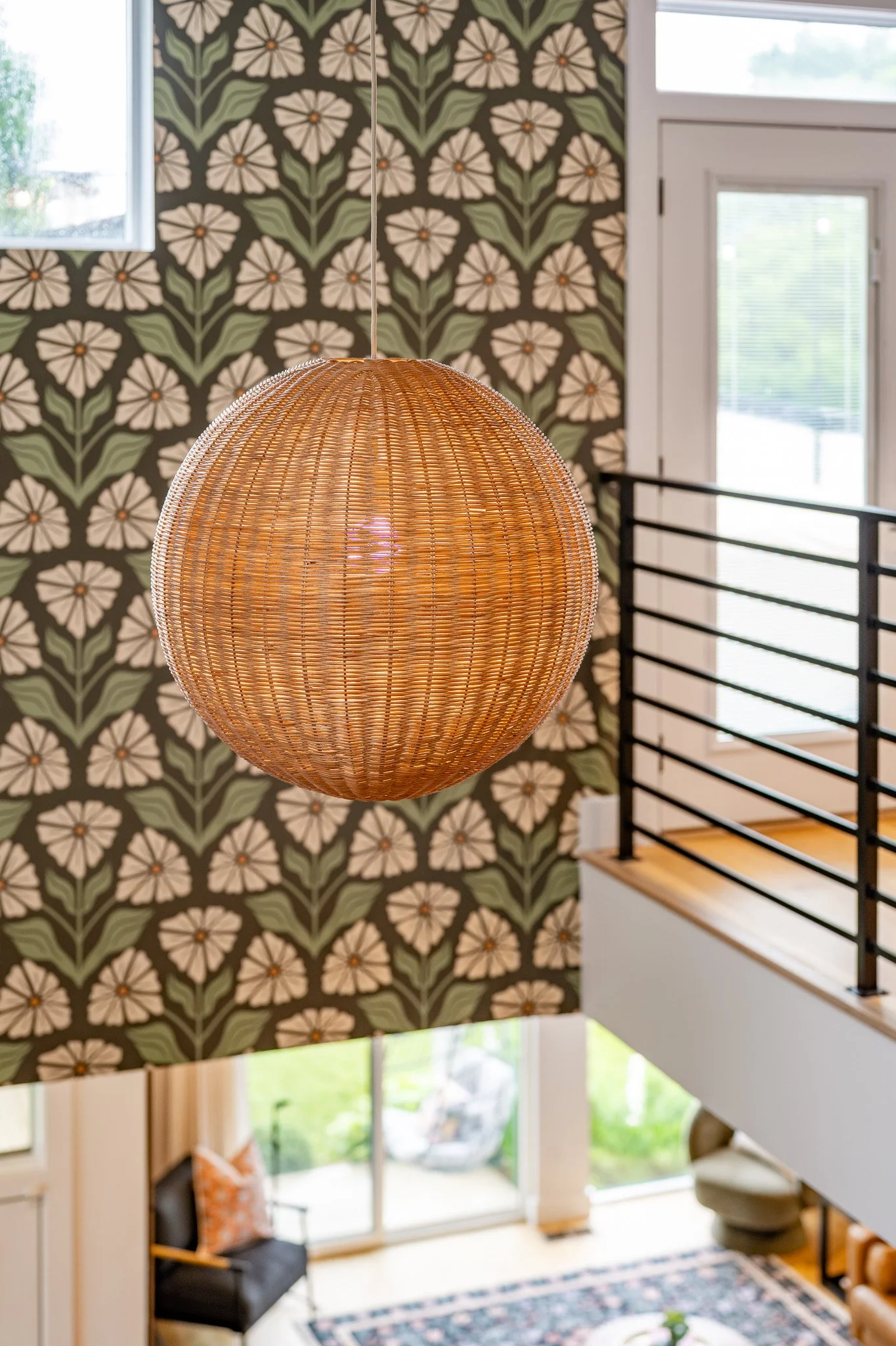 A round woven pendant light hanging from the ceiling of a room with a large window and floral wallpaper in green, white, and orange colors.
