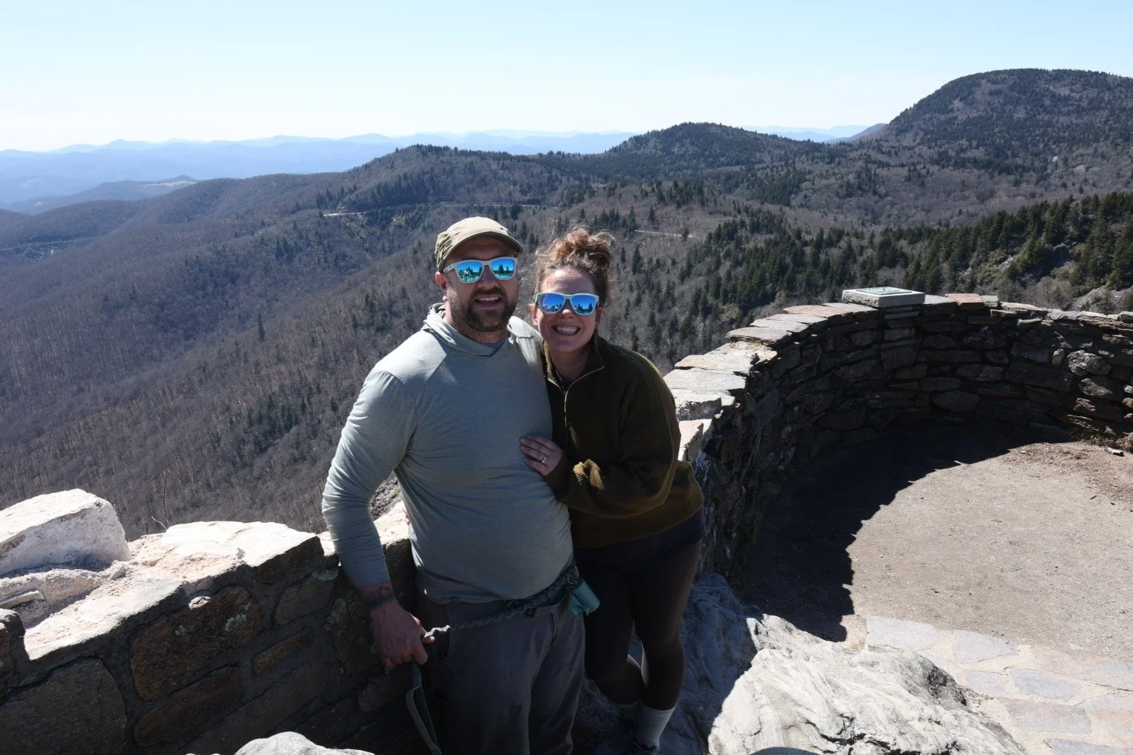 A smiling man and woman wearing sunglasses at a mountain viewpoint with a stone railing, overlooking a forested mountain landscape on a clear day.