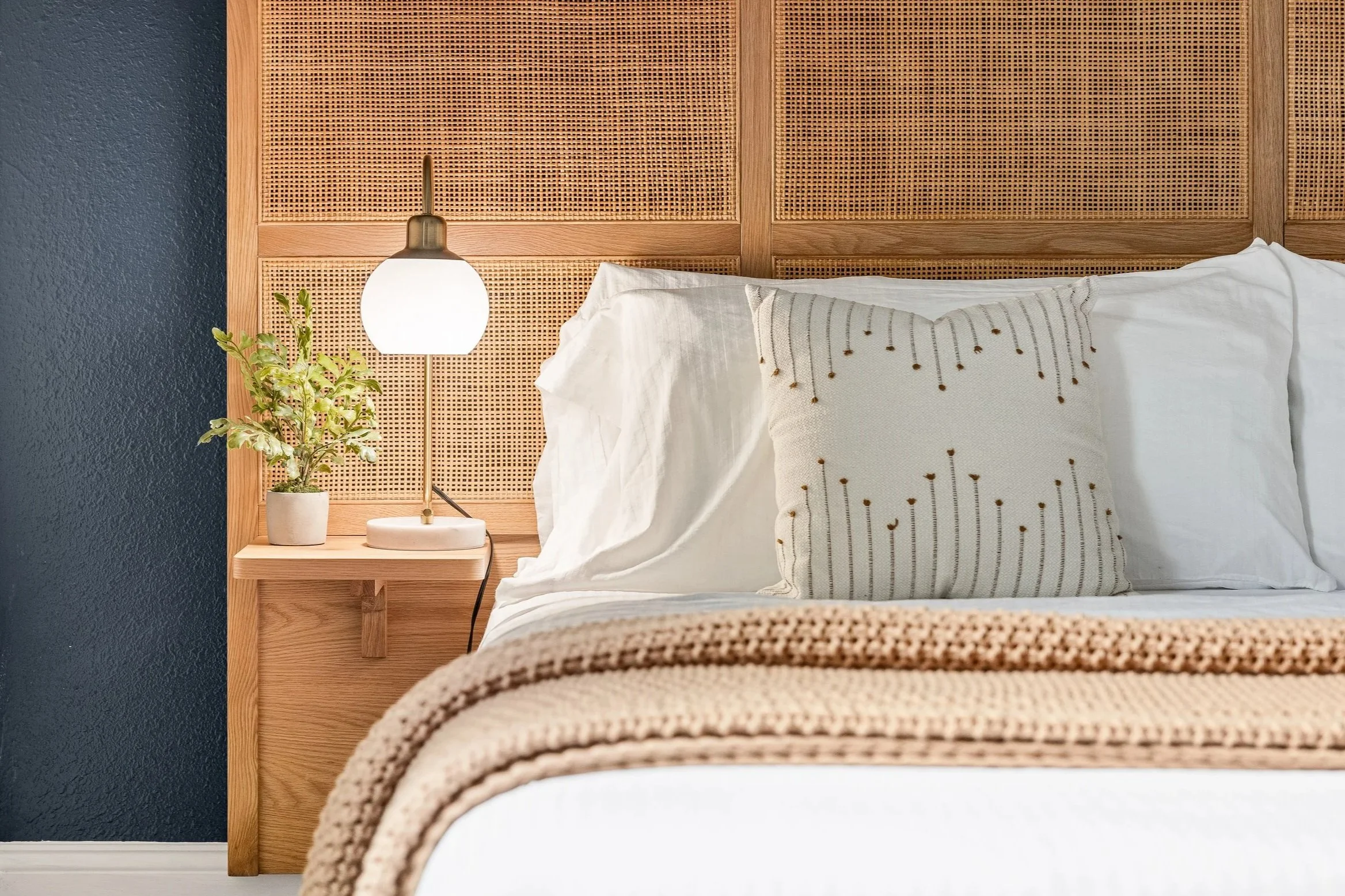 A neatly made bed with white bedding and a decorative pillow with dotted lines, a bedside table with a small potted plant, a modern white lamp, and a woven throw, all against a textured dark blue wall and a wooden headboard with woven cane panels.