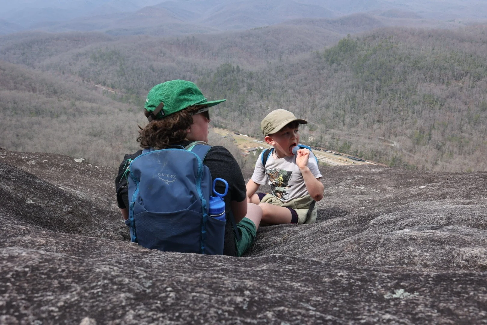 Two children sitting on a large rock overlooking a mountainous landscape, one boy eating a popsicle, both wearing backpacks and hats.