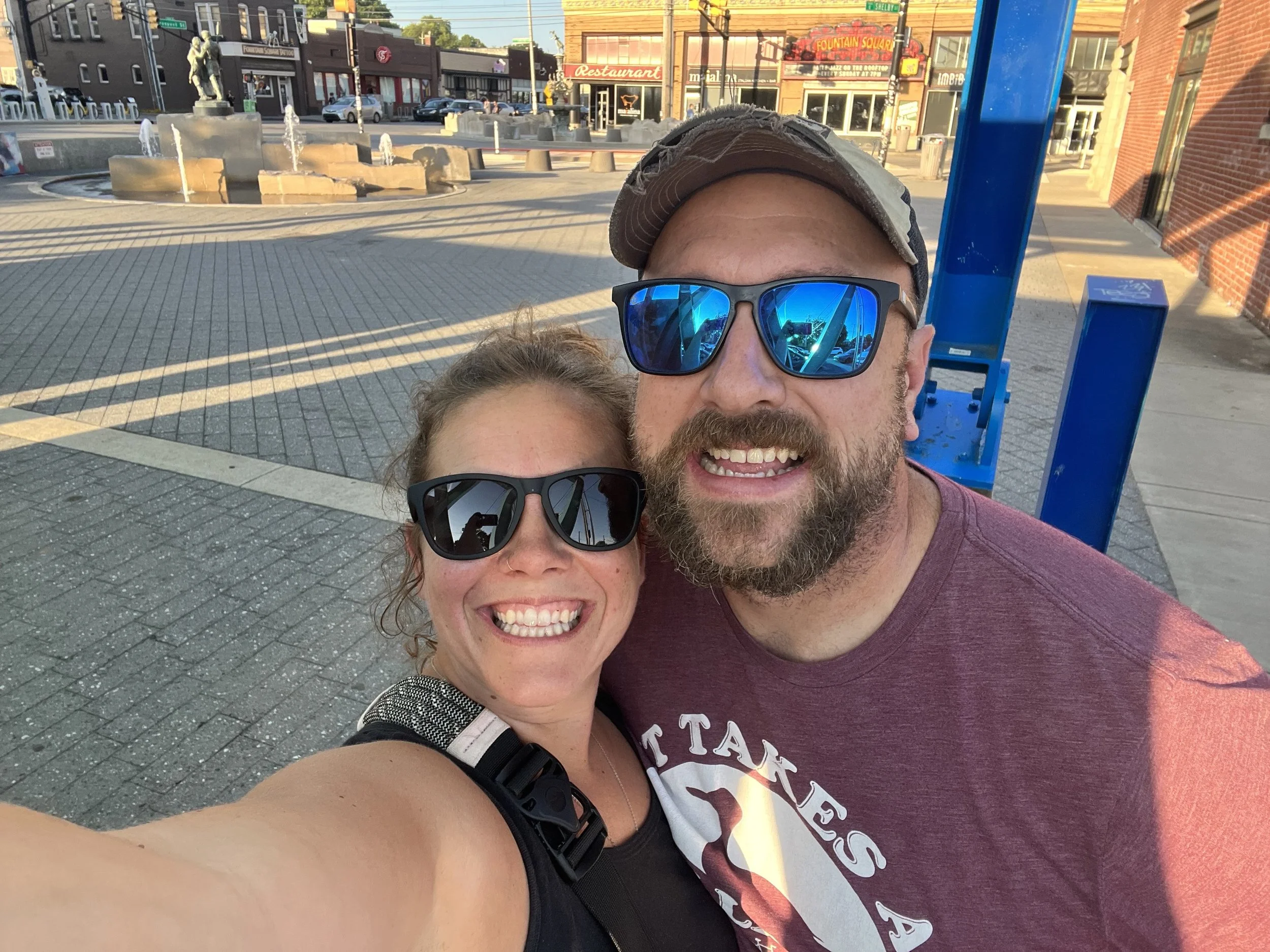 A man and woman taking a selfie outdoors at a city square. The man wears a baseball cap and sunglasses, and the woman is next to him wearing sunglasses and a black top. The background shows a fountain, buildings, and storefronts.