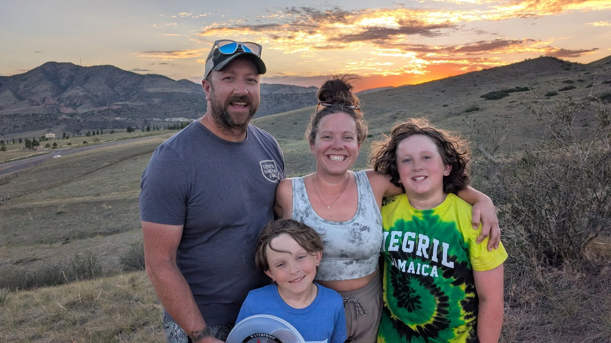 A group of five people standing outdoors during sunset on a hillside with mountains in the background. They are smiling and appear to be enjoying a family outing.