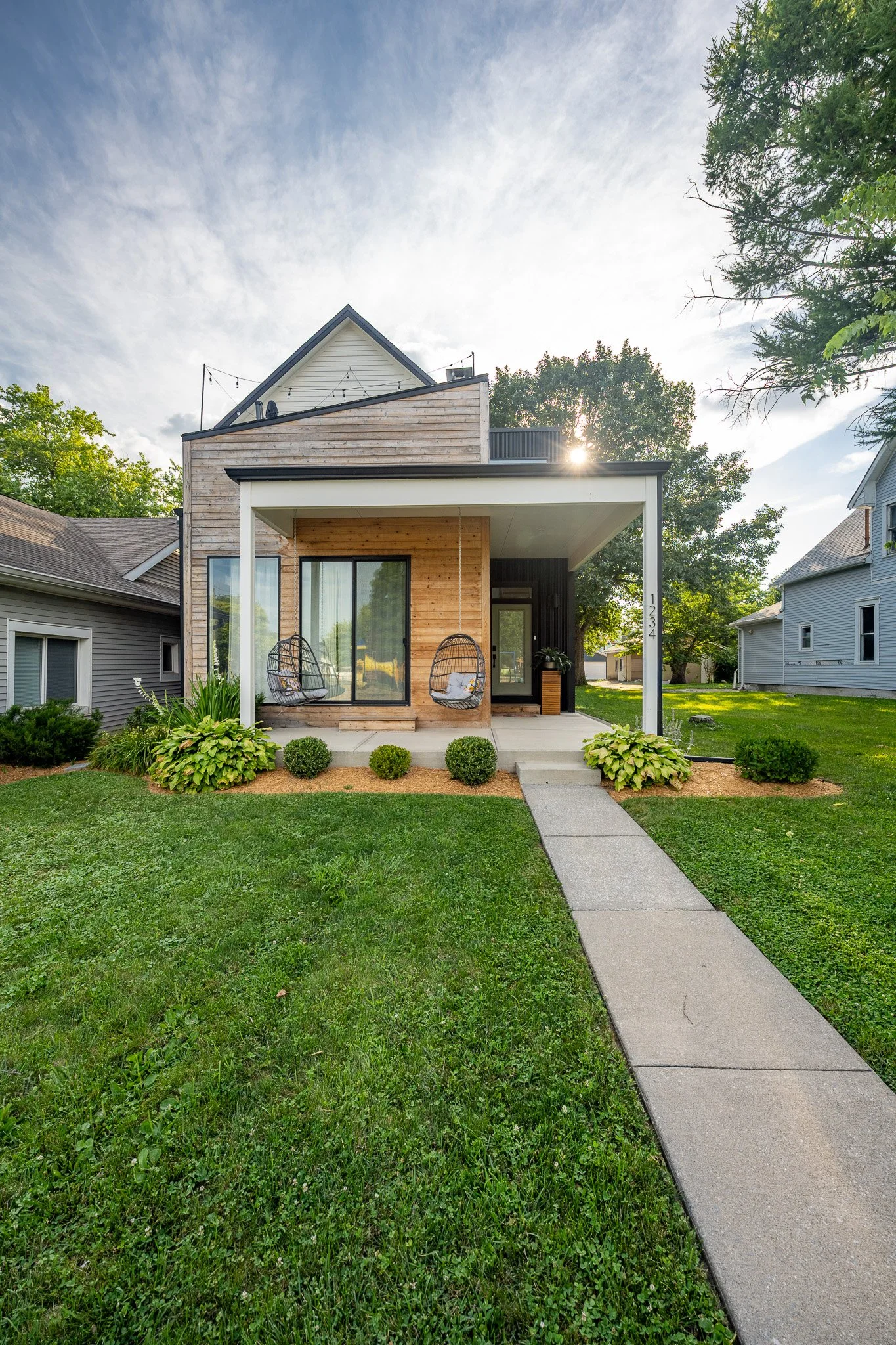 Front view of a modern small house with a porch, swing chairs, and landscaped garden, surrounded by other houses and trees, under a partly cloudy sky with sunlight.